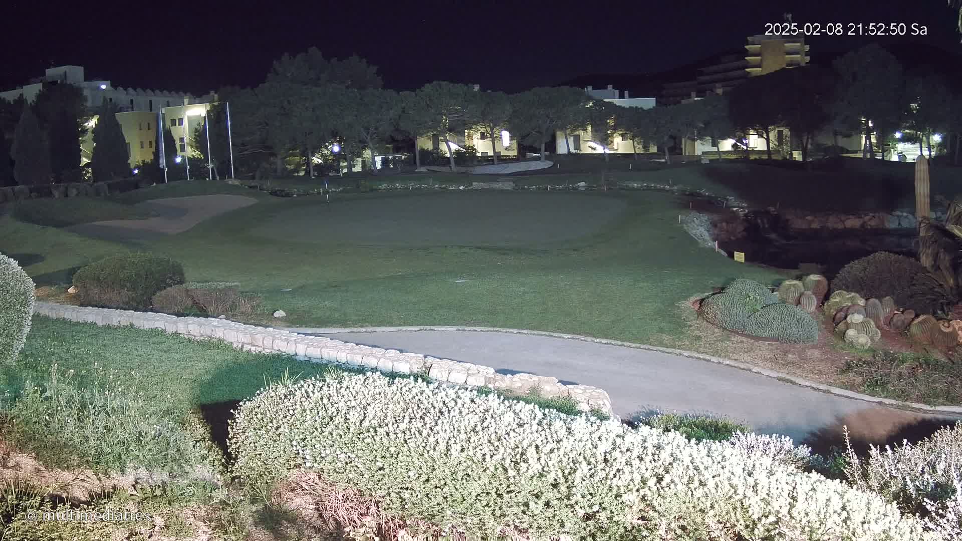 A nighttime view of a putting green on a golf course, lit by artificial light, with a building and trees in the background.