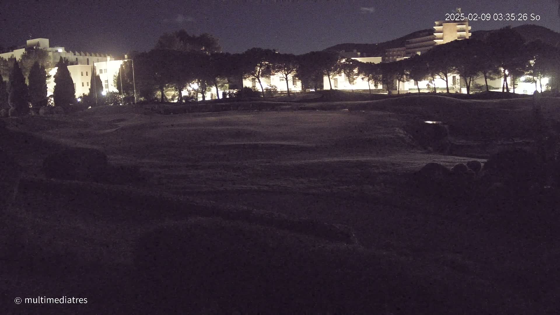 A dark, nighttime scene shows a line of lit buildings behind a row of trees and a large, dark, empty field.