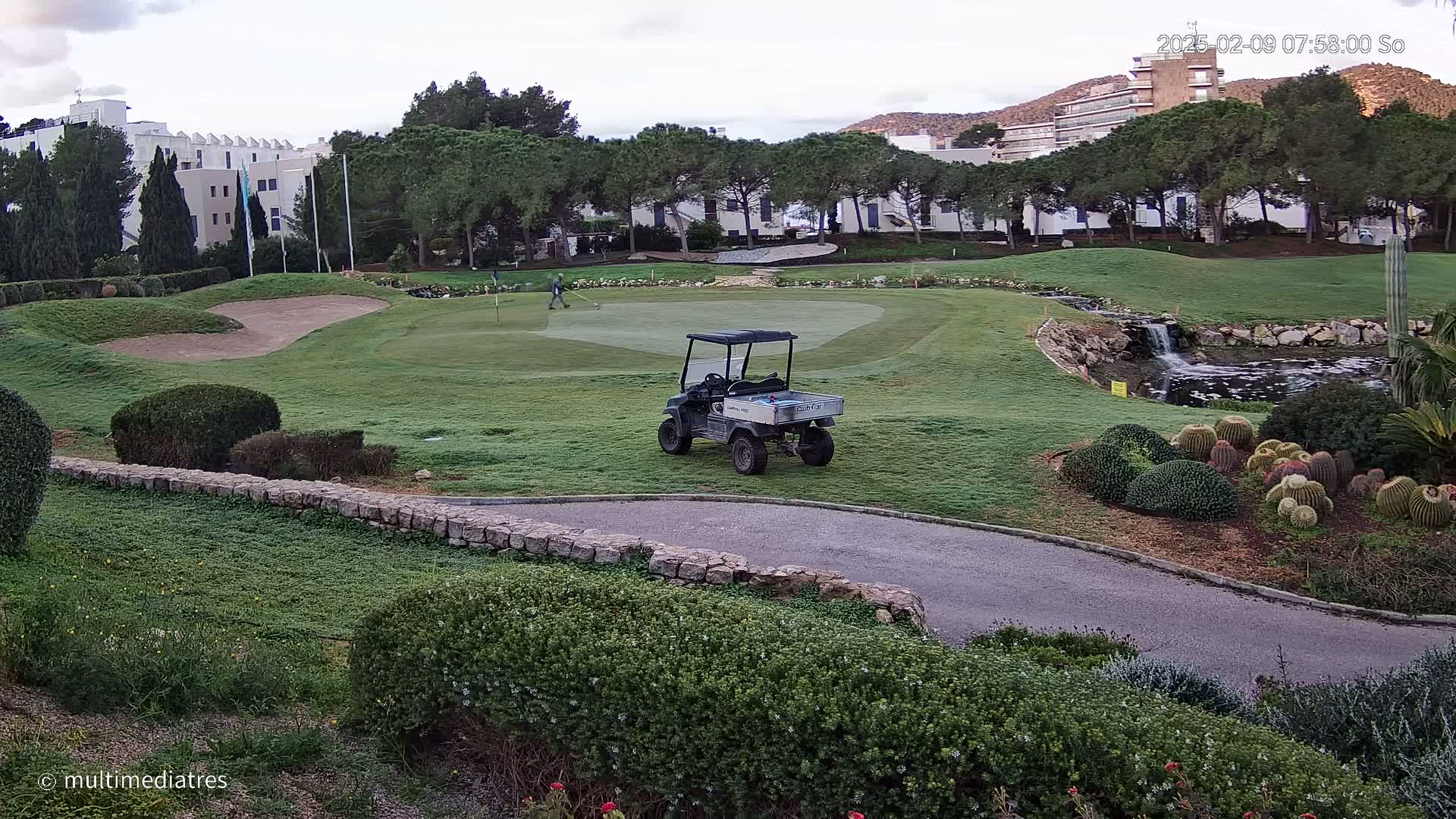 A golf course with a golf cart parked near a putting green, a small waterfall, and a person tending the course under an overcast sky.