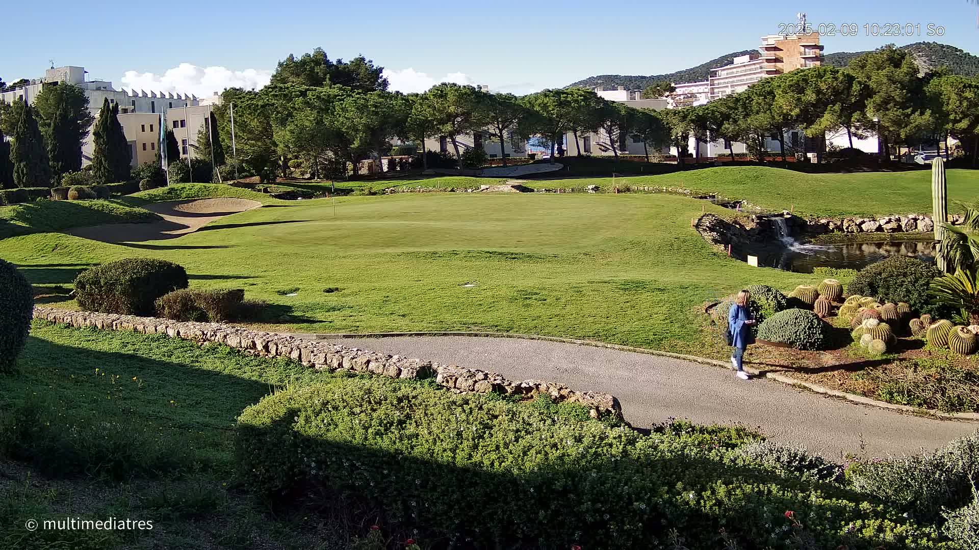 A woman walks along a path on a sunny day next to a golf course with a small pond and cacti.