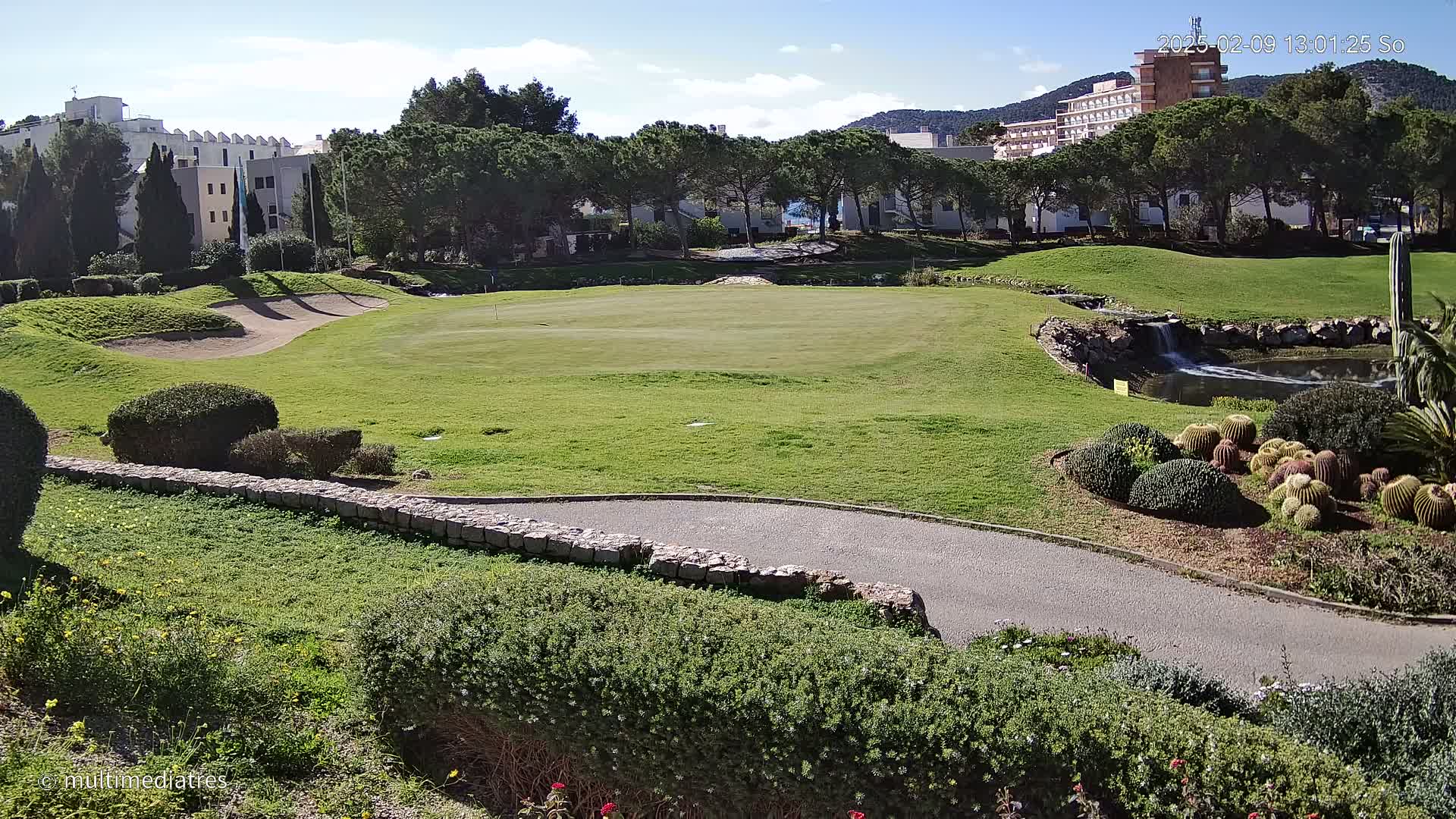 A golf course with a green, sand trap, and a small pond with a waterfall is situated near buildings under a clear, sunny sky.