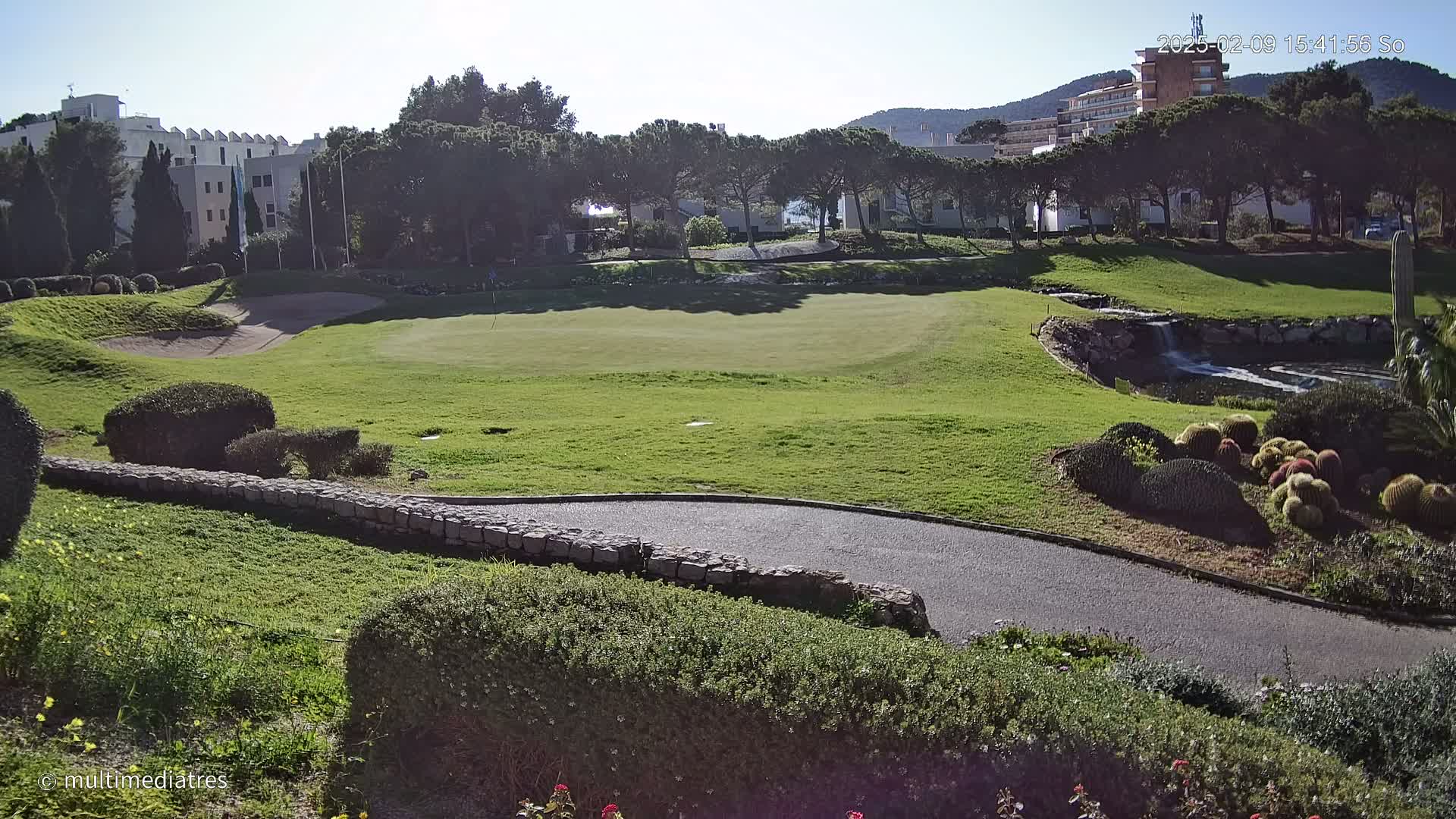 A sunny view of a golf course putting green, bordered by manicured lawns, a stone wall, and a small waterfall, with buildings and trees in the background.