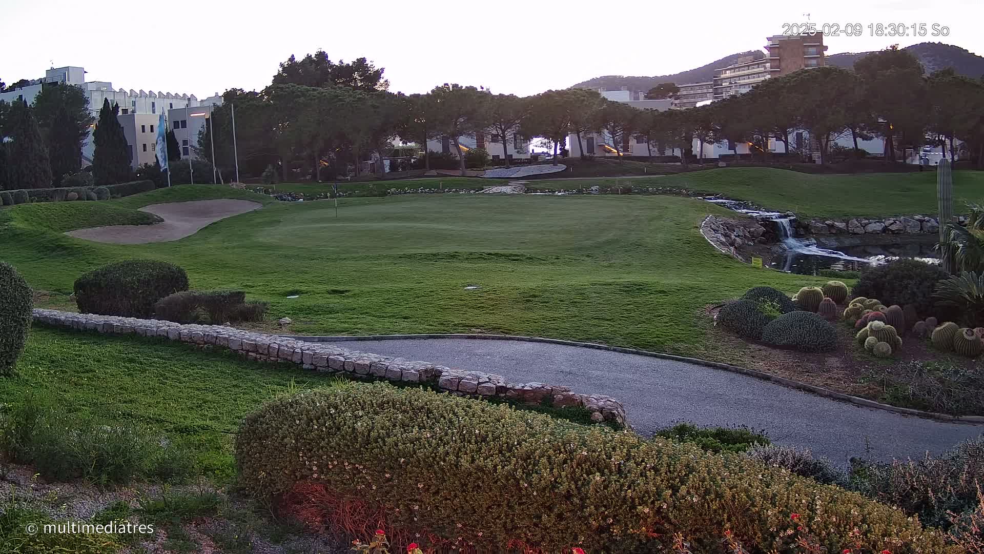 A golf course green with a sand trap, a small waterfall, and several cacti in the foreground, all under a twilight sky.