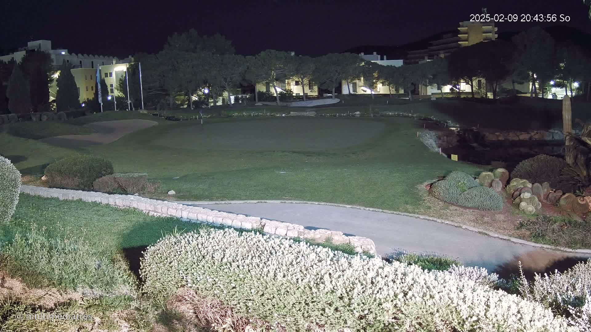 A nighttime view of a putting green and surrounding landscaping, illuminated by artificial lights, near a building complex.