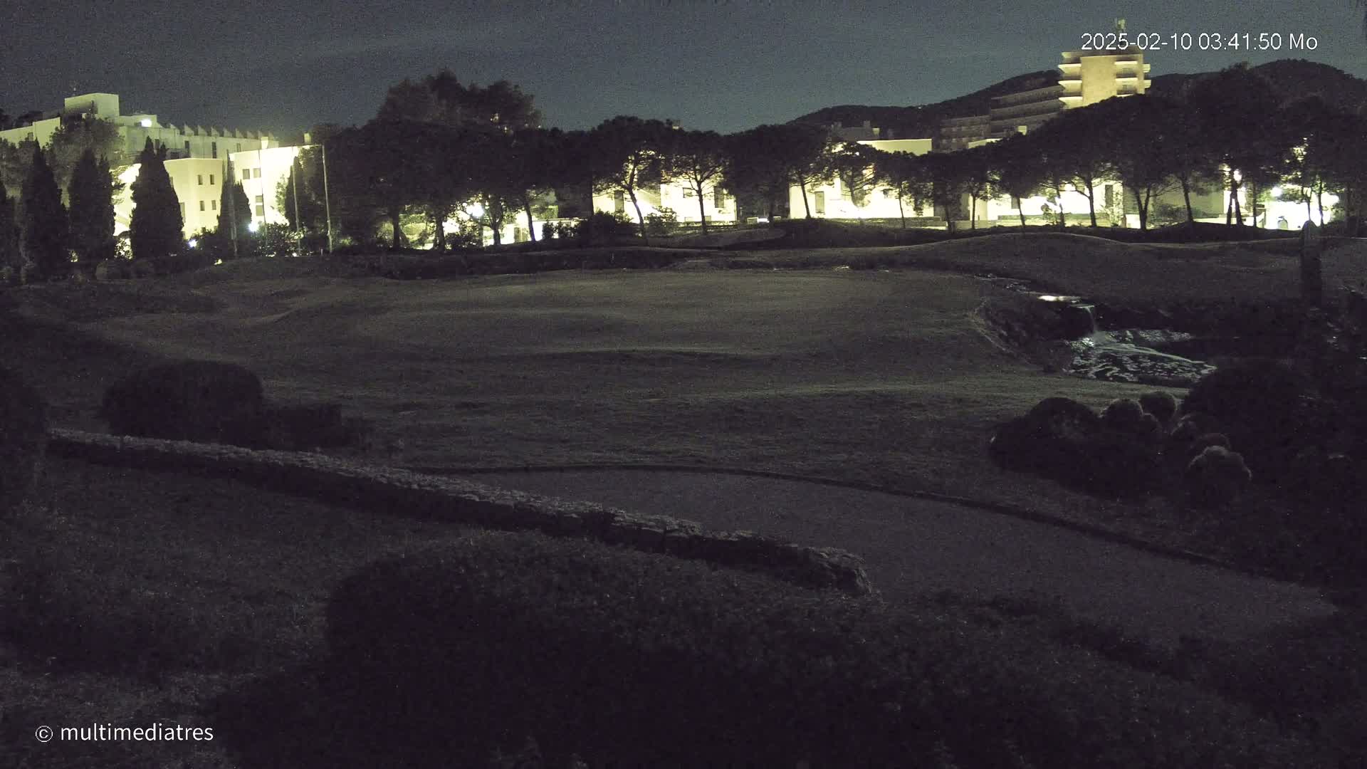A nighttime view of a grassy area, possibly a golf course, with a low-lying water feature and illuminated buildings in the background.