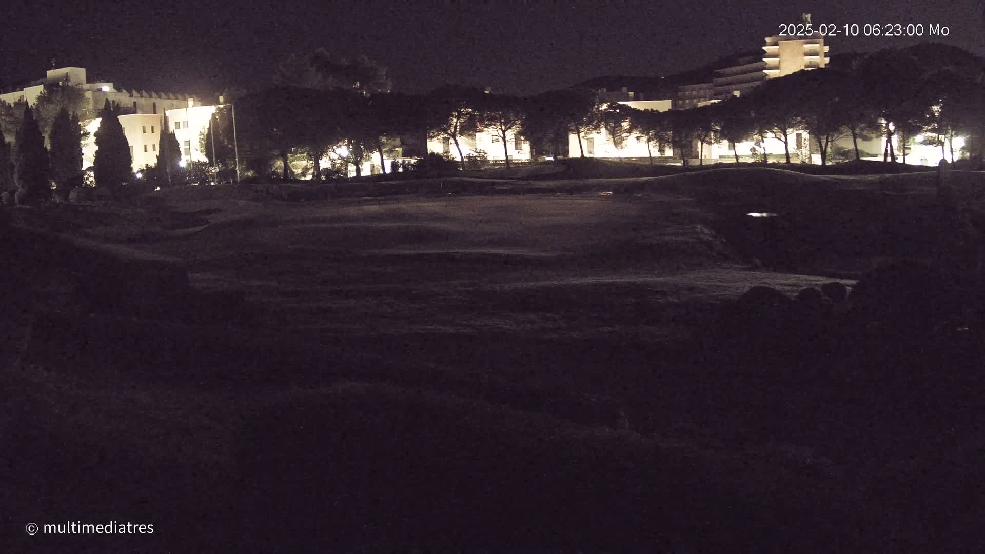 At night, illuminated buildings are partially visible behind a dark, grassy area.