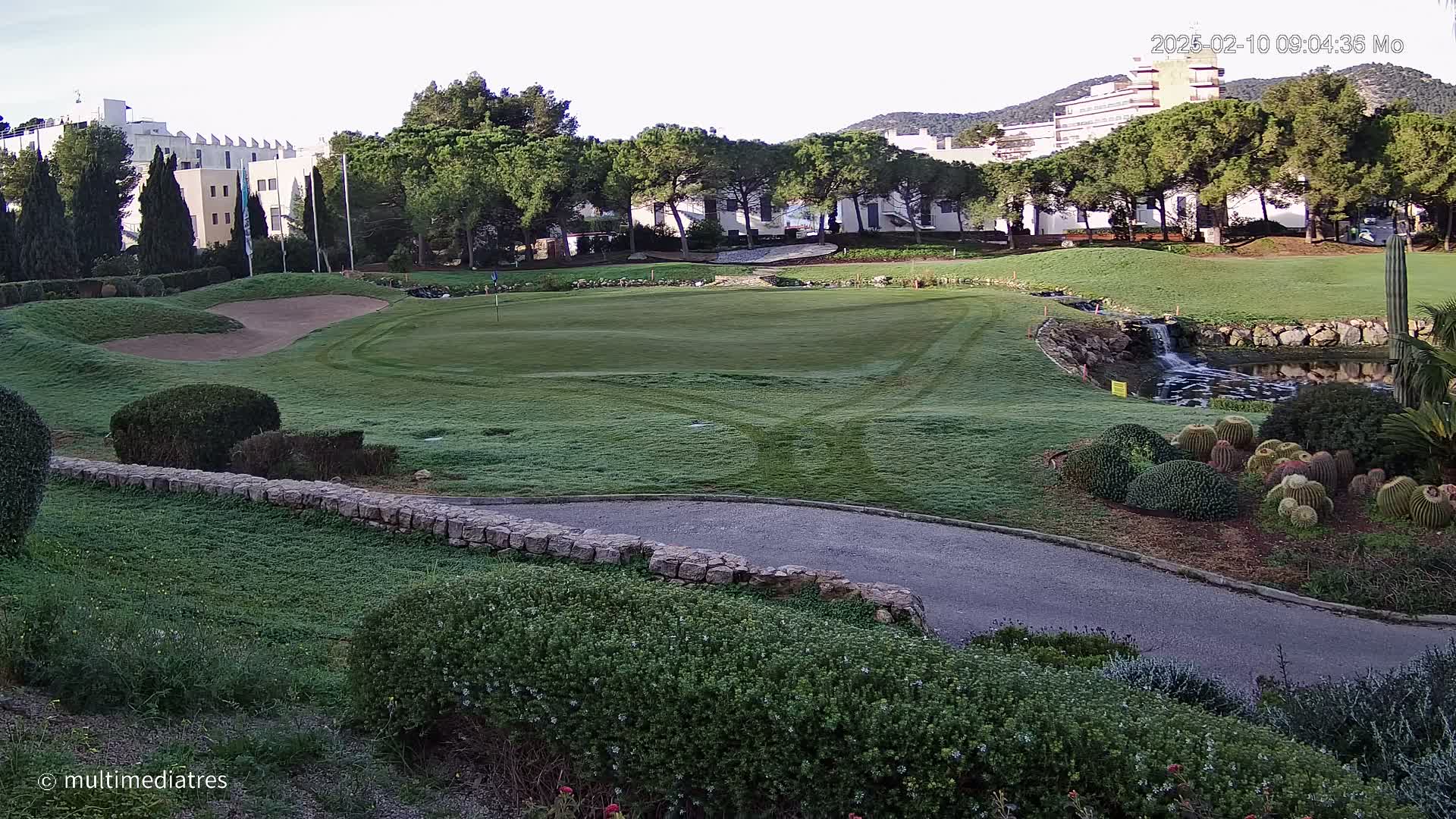 A golf course green with a sand trap, a small waterfall, and several cacti, all under a clear morning sky.