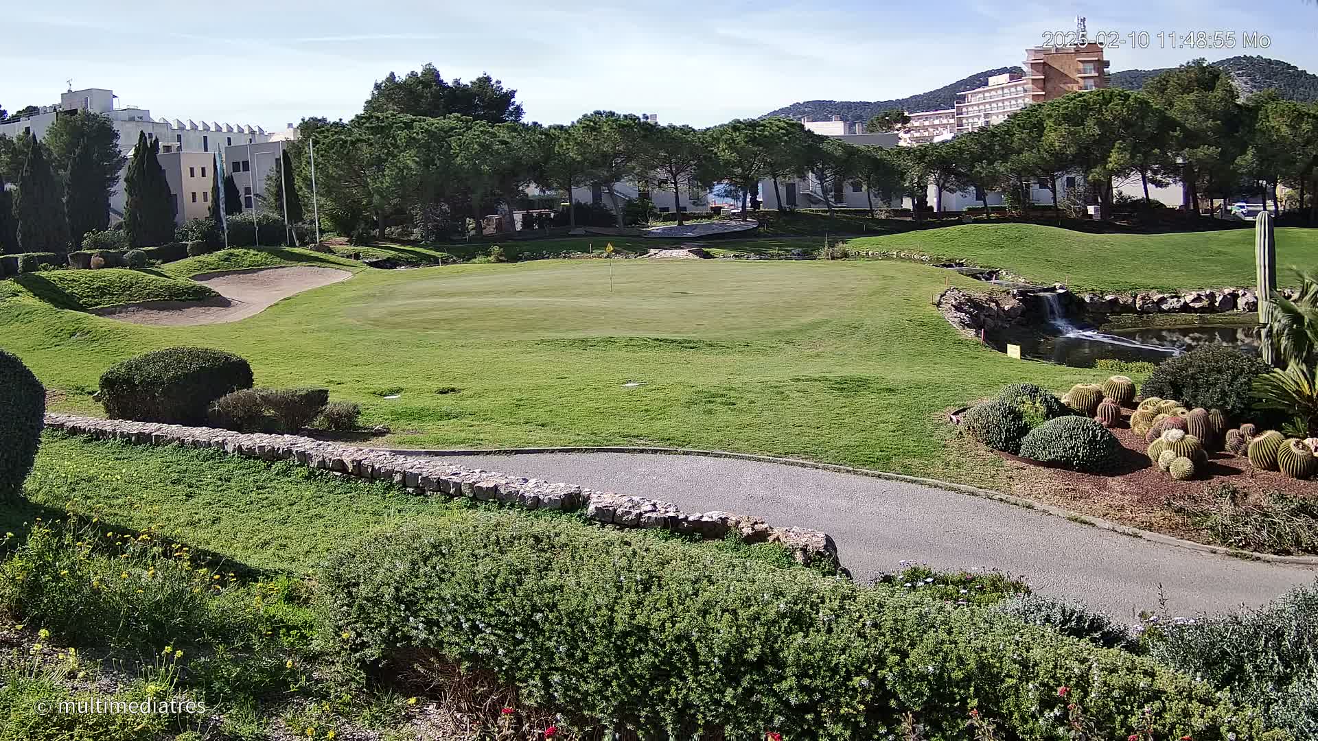 A golf course green with a sand trap, a small waterfall, and a collection of cacti is visible under a sunny sky, with buildings and trees in the background.