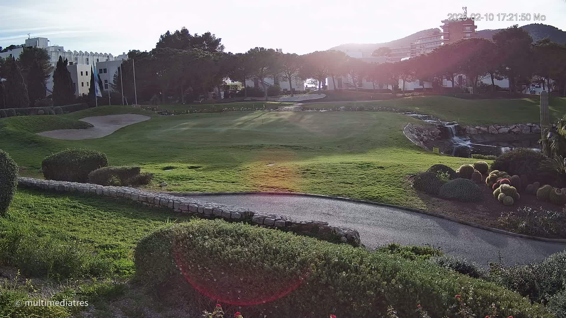 A golf course green with a sand trap and a small waterfall is bathed in the sunlight of a clear, sunny day, with buildings and trees in the background.
