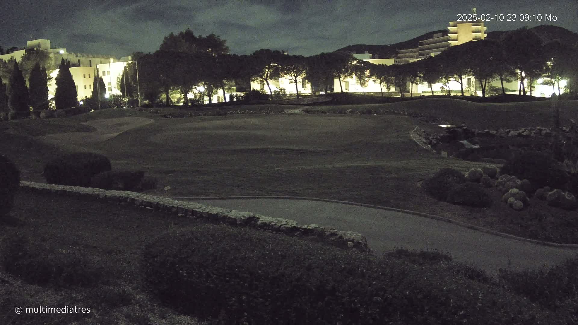 A nighttime view of a golf course with buildings illuminated in the background under a partly cloudy sky.