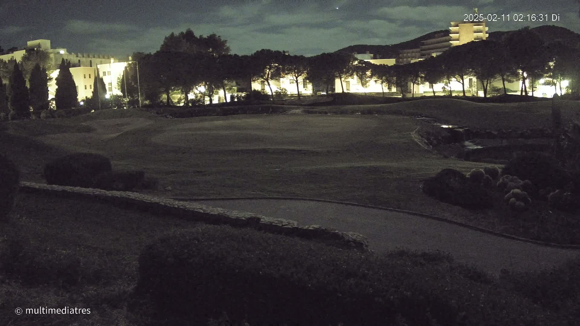 A nighttime view of a golf course with buildings and trees in the background under a partly cloudy sky.