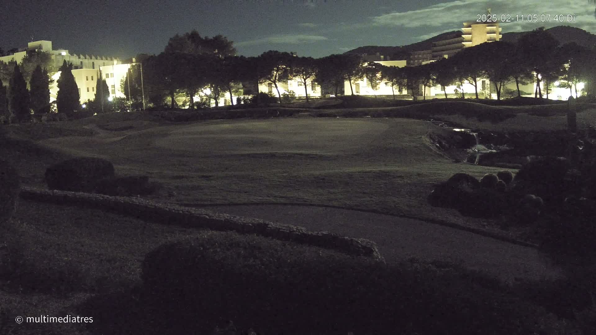 A nighttime view of a golf course with a building and trees in the background under a partly cloudy sky.