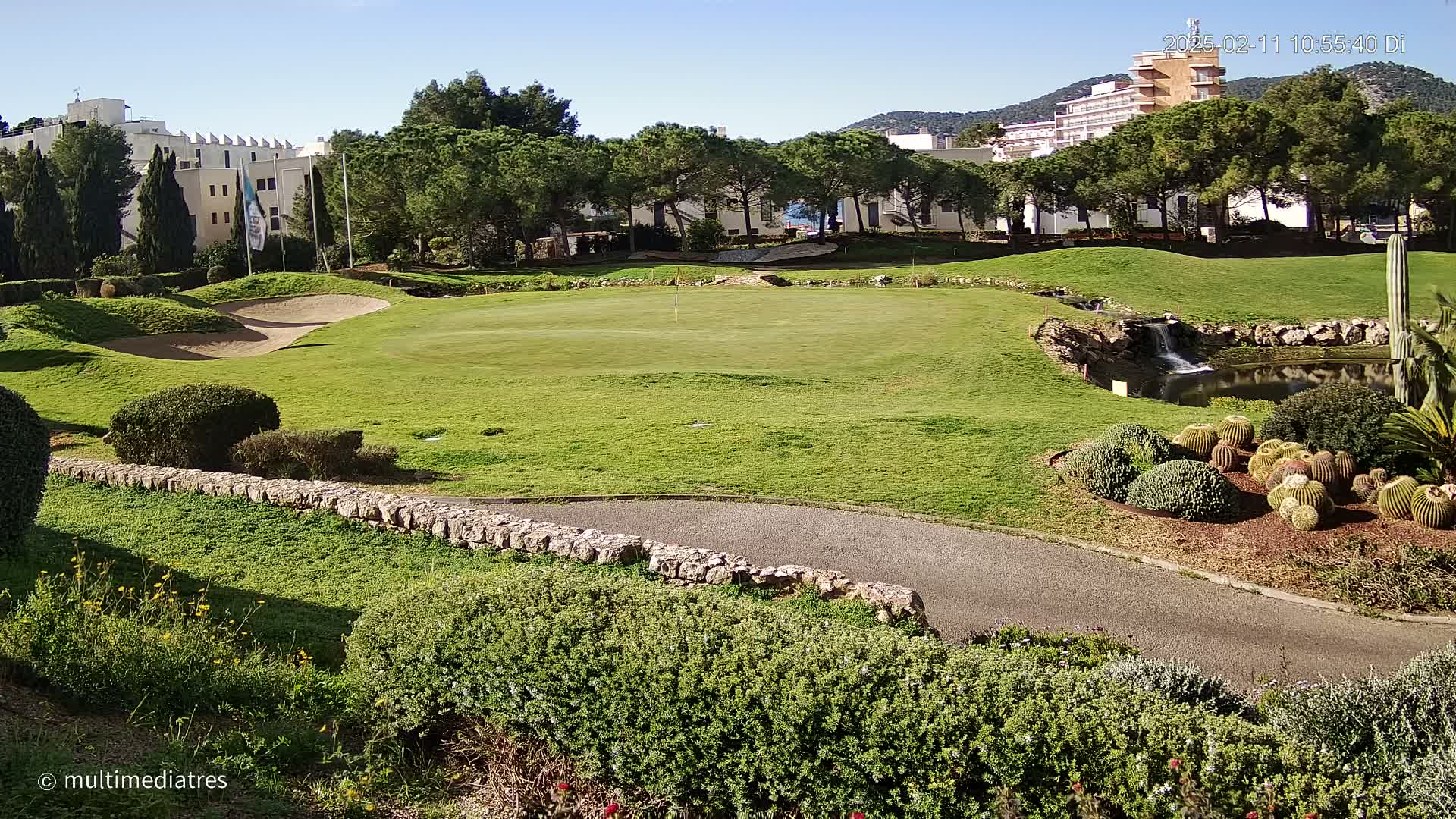 A sunny golf course with a sand trap, a small pond with a waterfall, and a backdrop of buildings and trees.