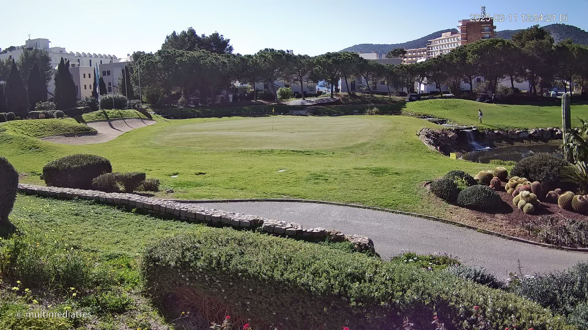 A sunny day reveals a golf course with a green, sand trap, a small waterfall, a pond, and various landscaping features, all set against a backdrop of buildings and trees.