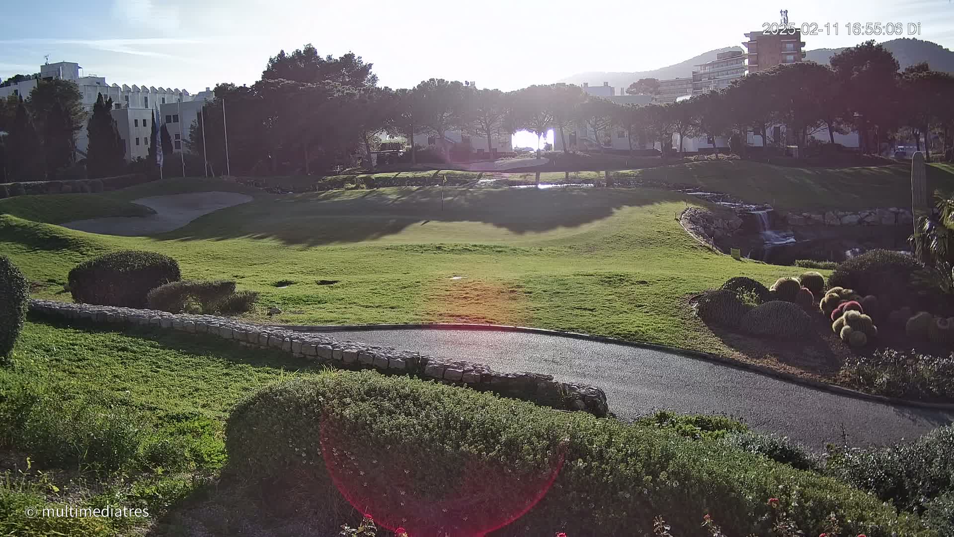 A sunny day on a golf course with a paved path winding through landscaped areas, near buildings and a small waterfall.