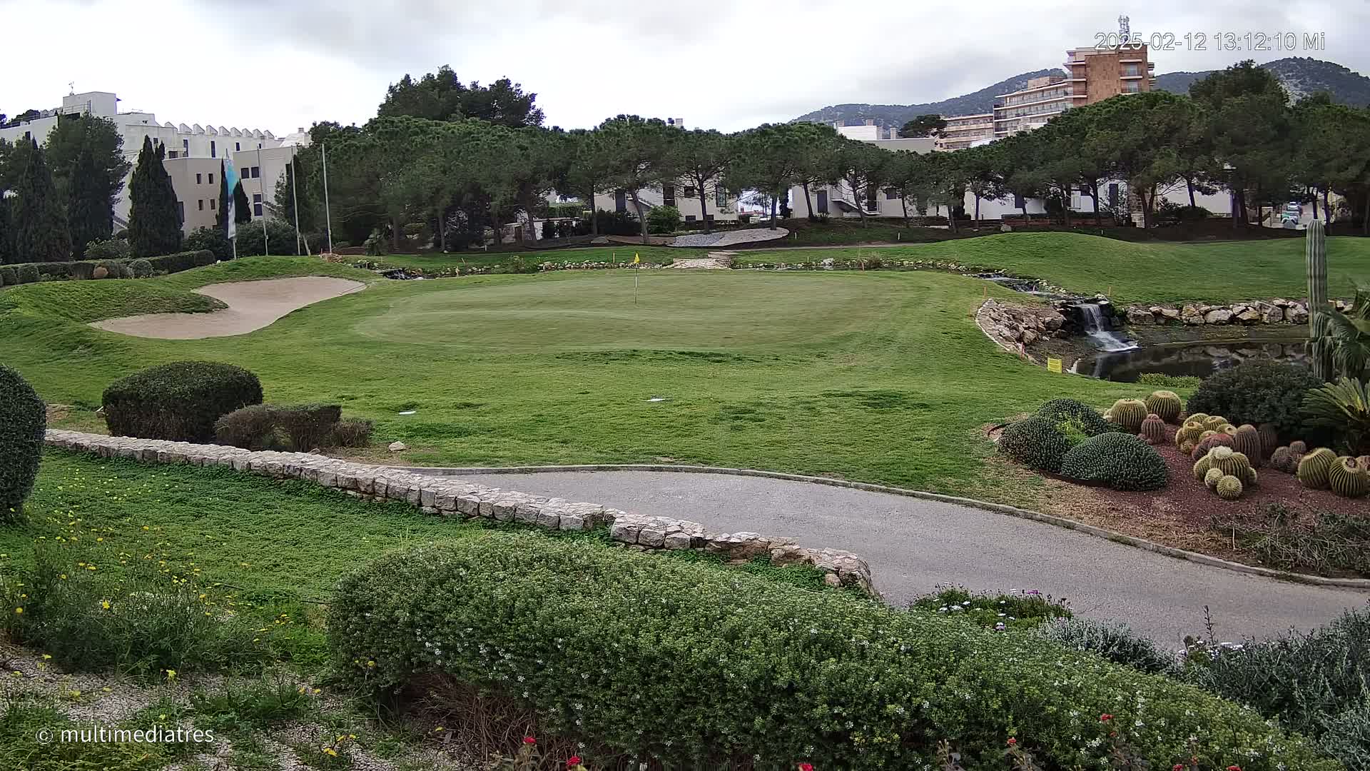 A golf course green with a sand trap, a small waterfall, and cacti, under an overcast sky.