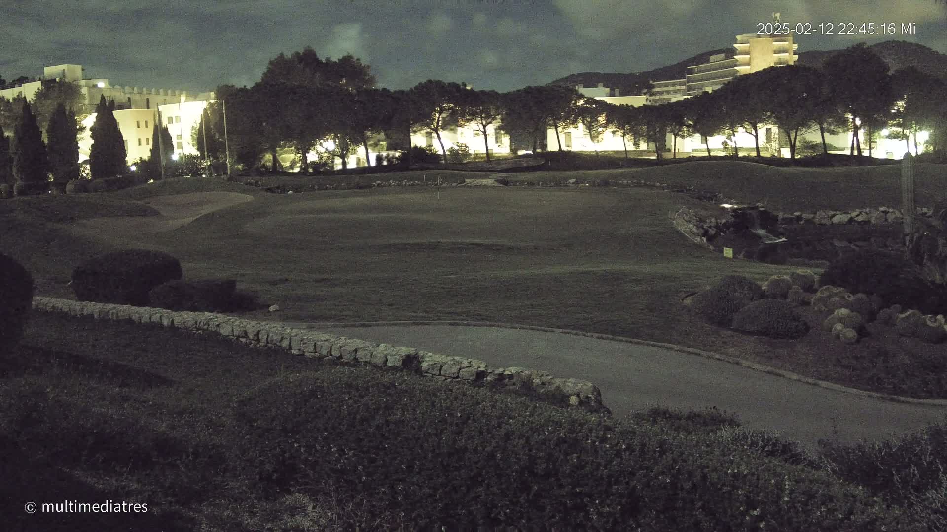 A nighttime view of a golf course with a low stone wall, landscaping, and buildings illuminated in the background under a cloudy sky.