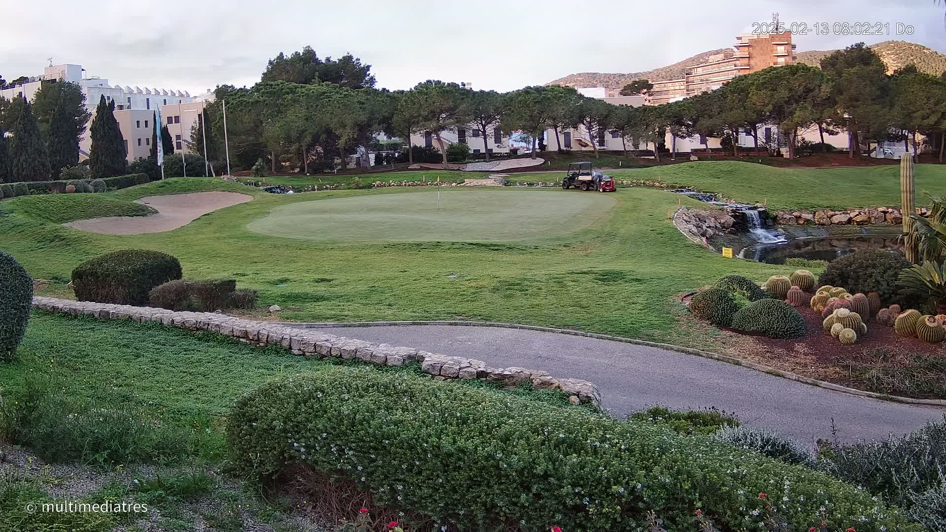 A golf course green with a sand trap, a small waterfall, and a variety of cacti and shrubs, situated near a multi-story building under a partly cloudy sky.