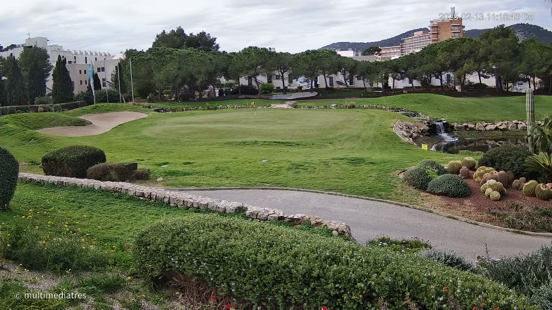 A golf course green with a sand trap, a small waterfall, and a collection of cacti is visible under an overcast sky.