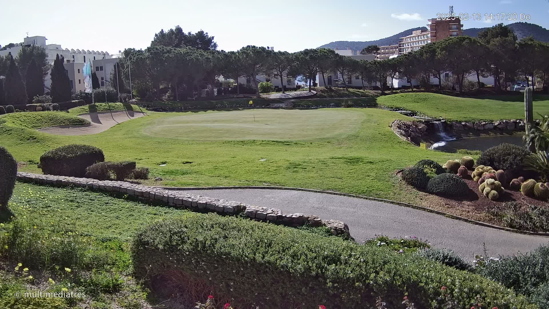 A sunny day reveals a golf course green with a sand trap, a small waterfall, and a variety of cacti and shrubs, all situated near buildings and trees.