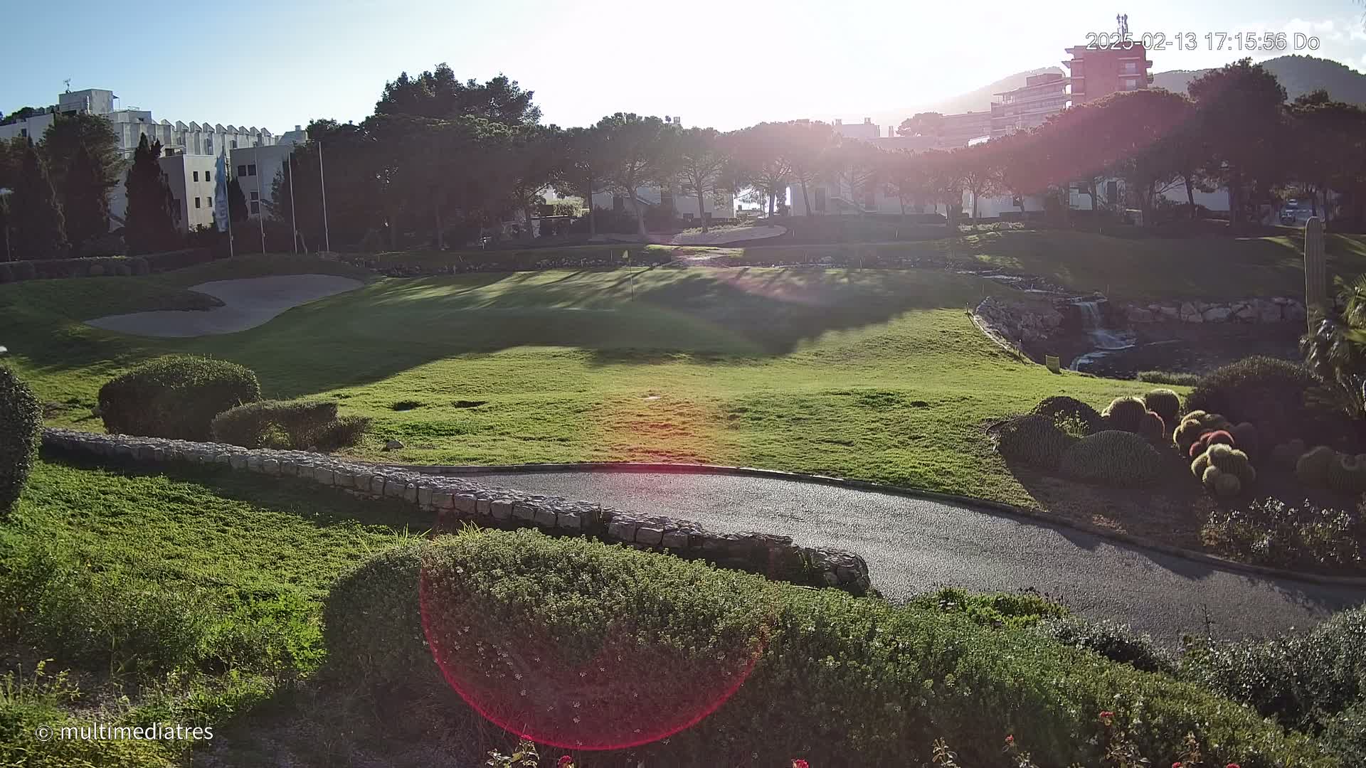 A sunny view of a golf course with a paved path winding through manicured lawns and landscaping, featuring a small waterfall and buildings in the background.