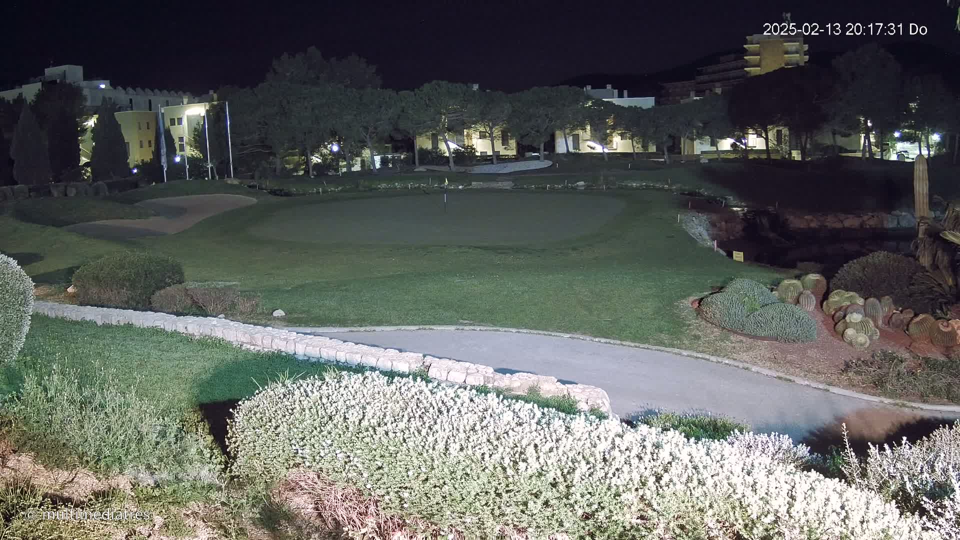 A nighttime view of a putting green at a golf course next to buildings and landscaping, including a low stone wall and flowering shrubs.