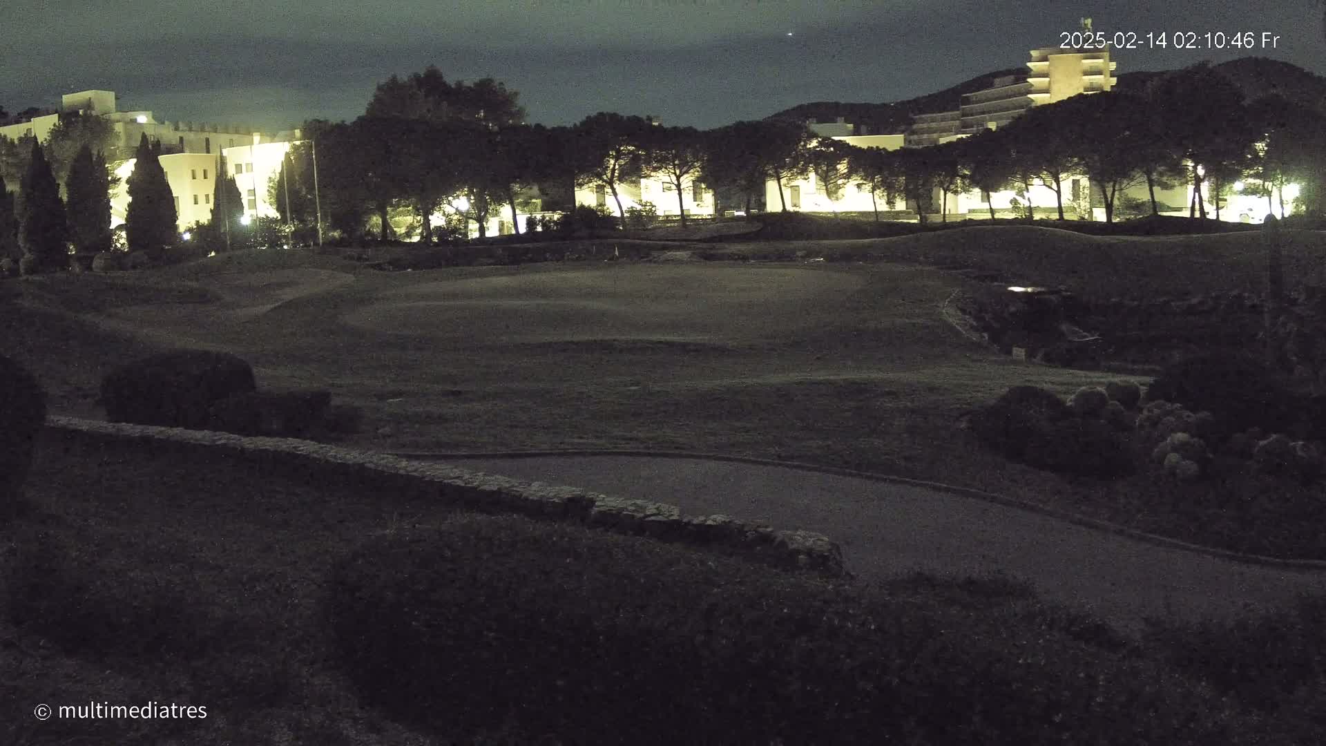 A nighttime view of a golf course with buildings in the background under a mostly clear sky.