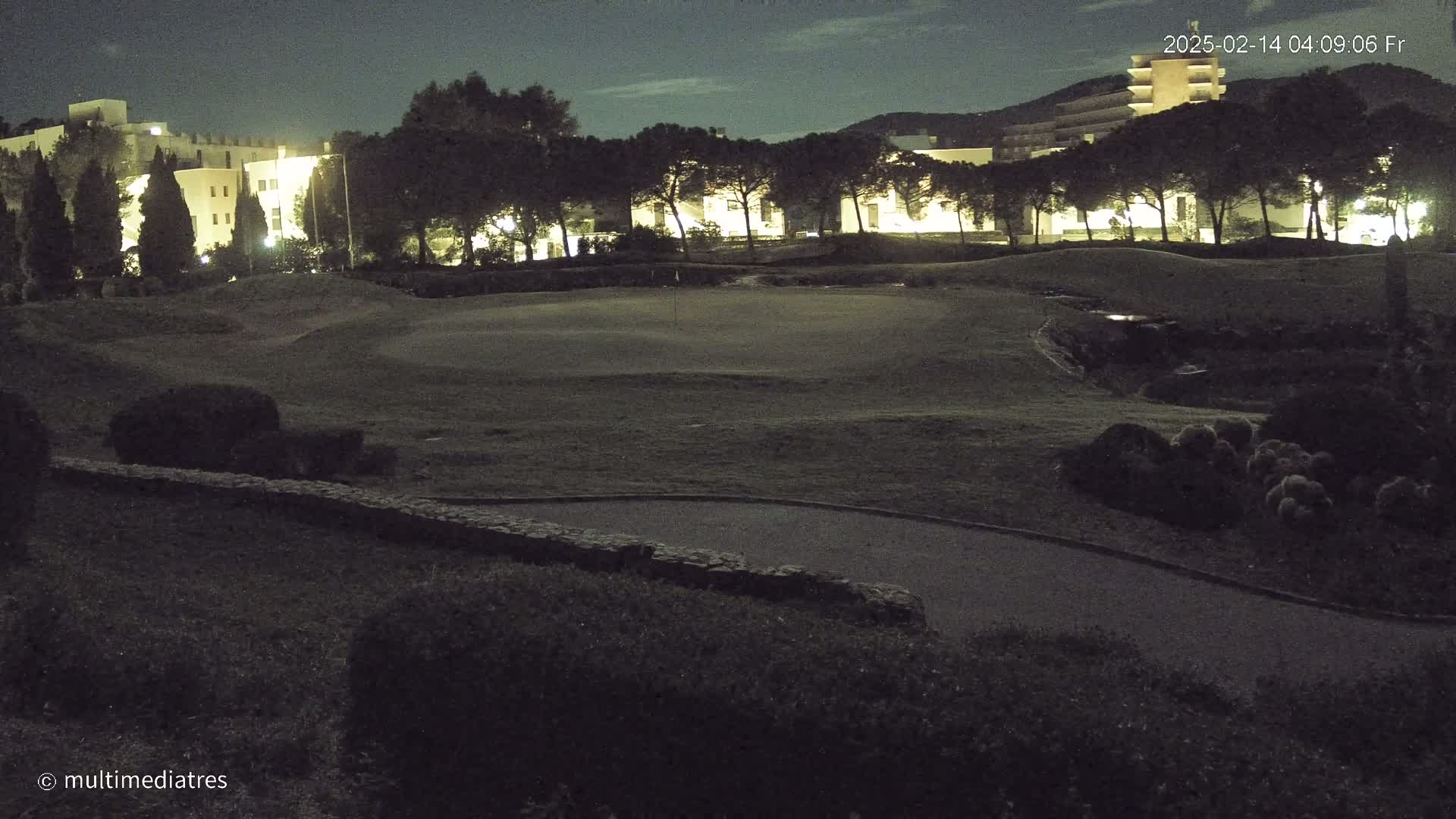 A nighttime view of a golf course with a building and trees illuminated in the background under a partly cloudy sky.