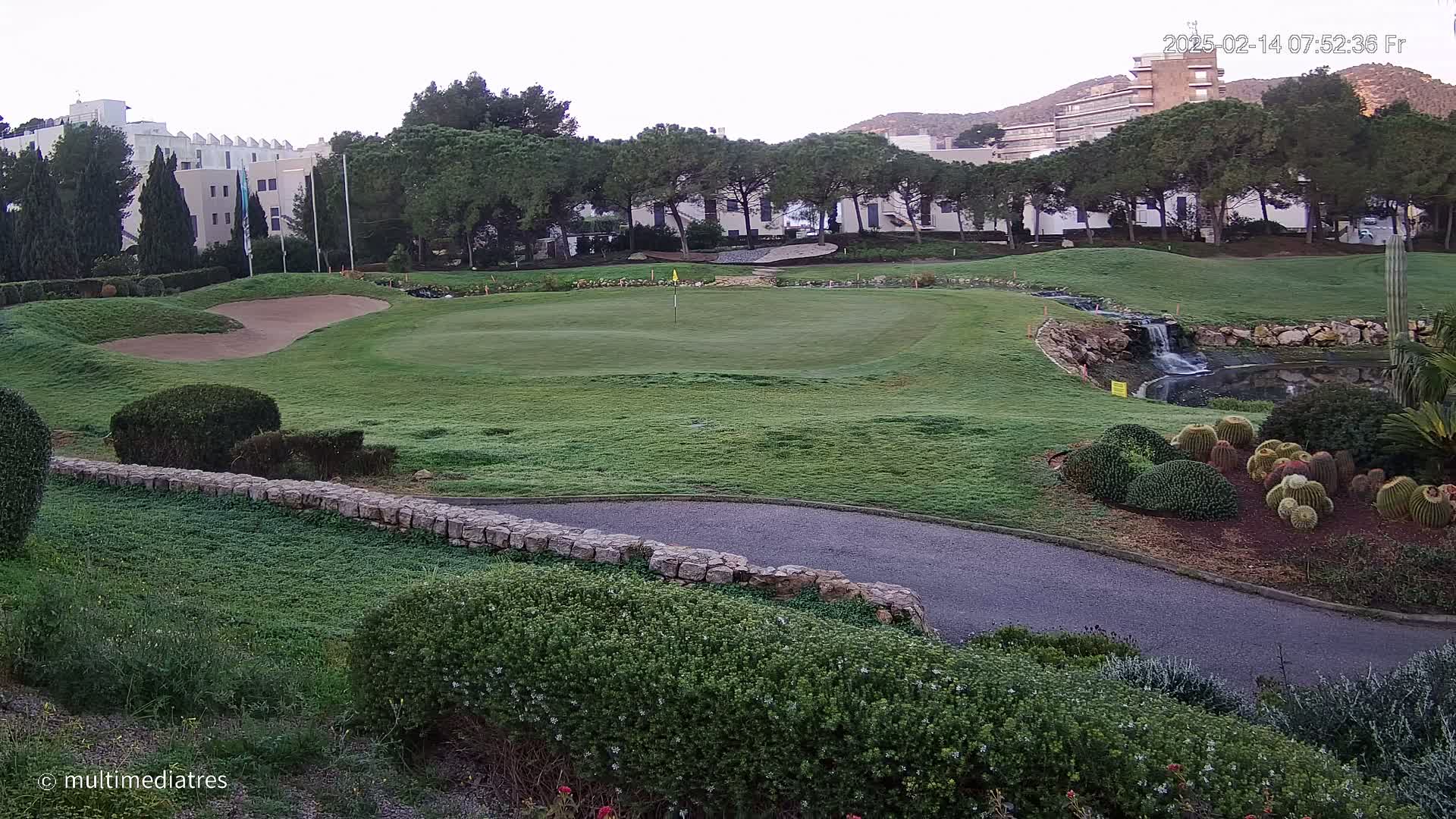 A golf course green with a sand trap, a small waterfall, and a variety of cacti and shrubs, all under a clear, early morning sky.