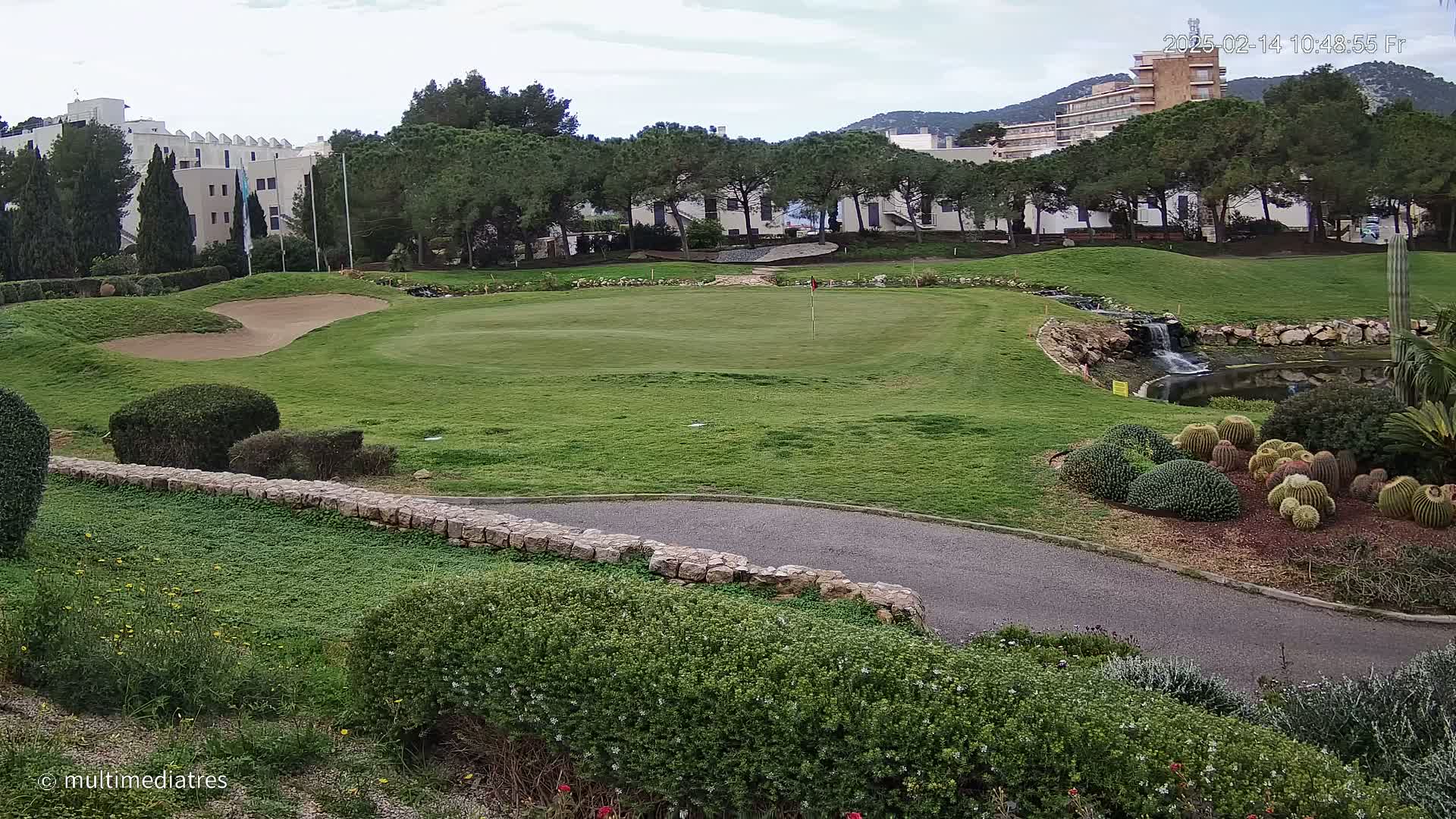A golf course green with a sand trap, a small waterfall, and cacti, all under a cloudy sky.