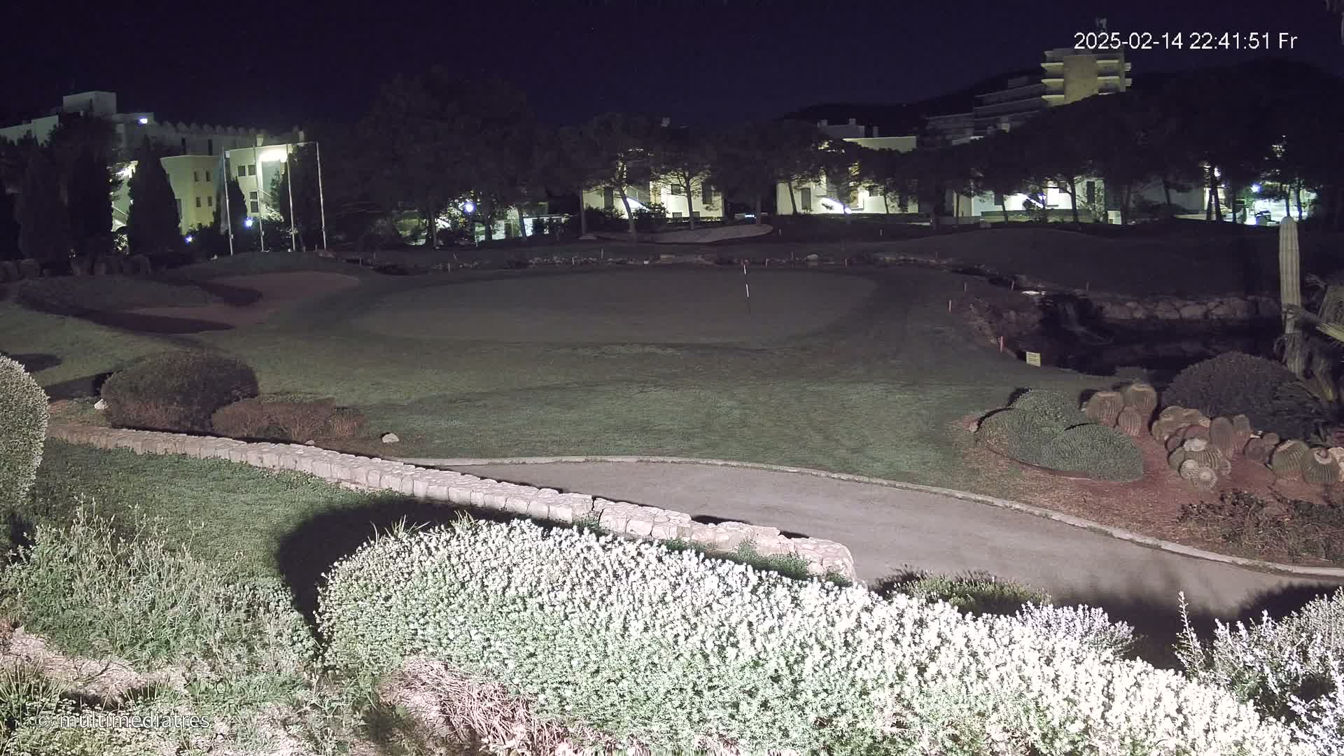 A nighttime view of a golf course green, bordered by shrubs and a low stone wall, with a building visible in the background under a clear, dark sky.