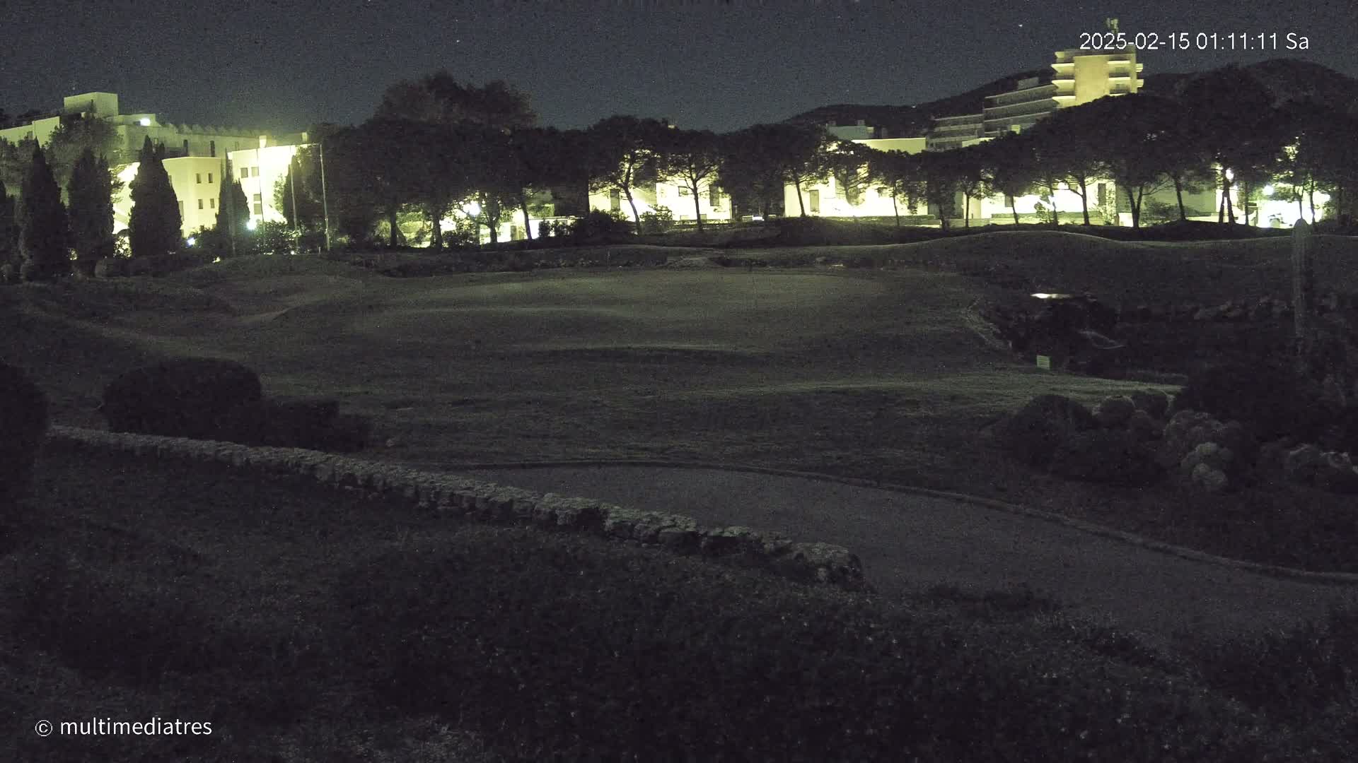 A nighttime view of a golf course with illuminated buildings in the background under a clear, starry sky.