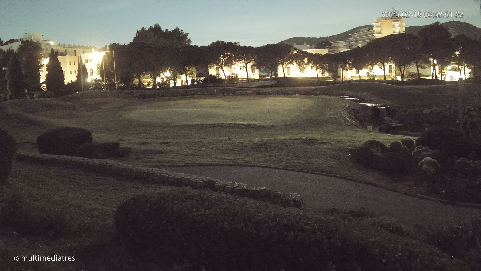 A nighttime view of a golf course with illuminated buildings and trees in the background under a dark sky.