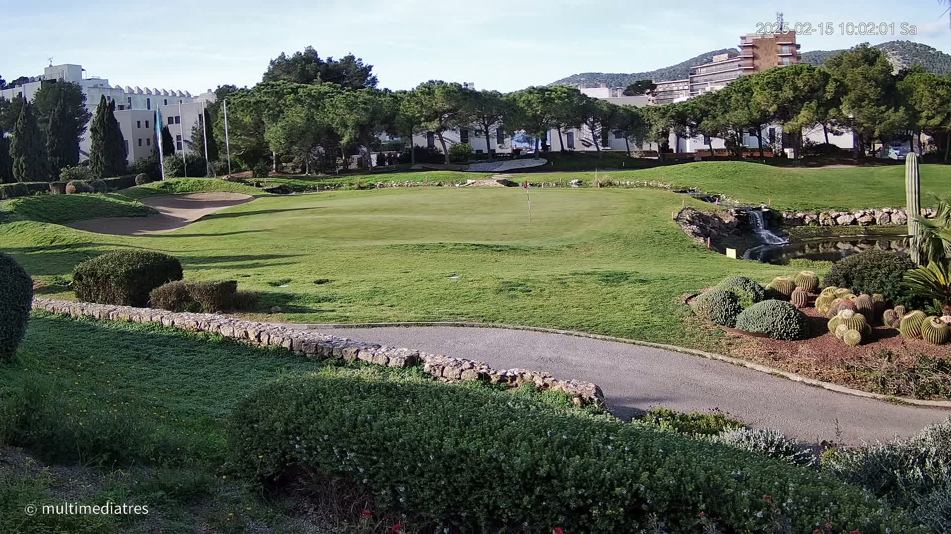 A sunny golf course with a small pond and cactus garden, situated near buildings and a mountain range.