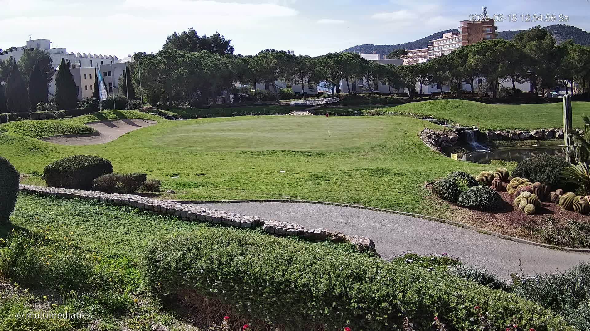 A sunny day on a golf course with a putting green, sand trap, small pond and waterfall, bordered by manicured shrubs and cactus gardens, with buildings and trees in the background.