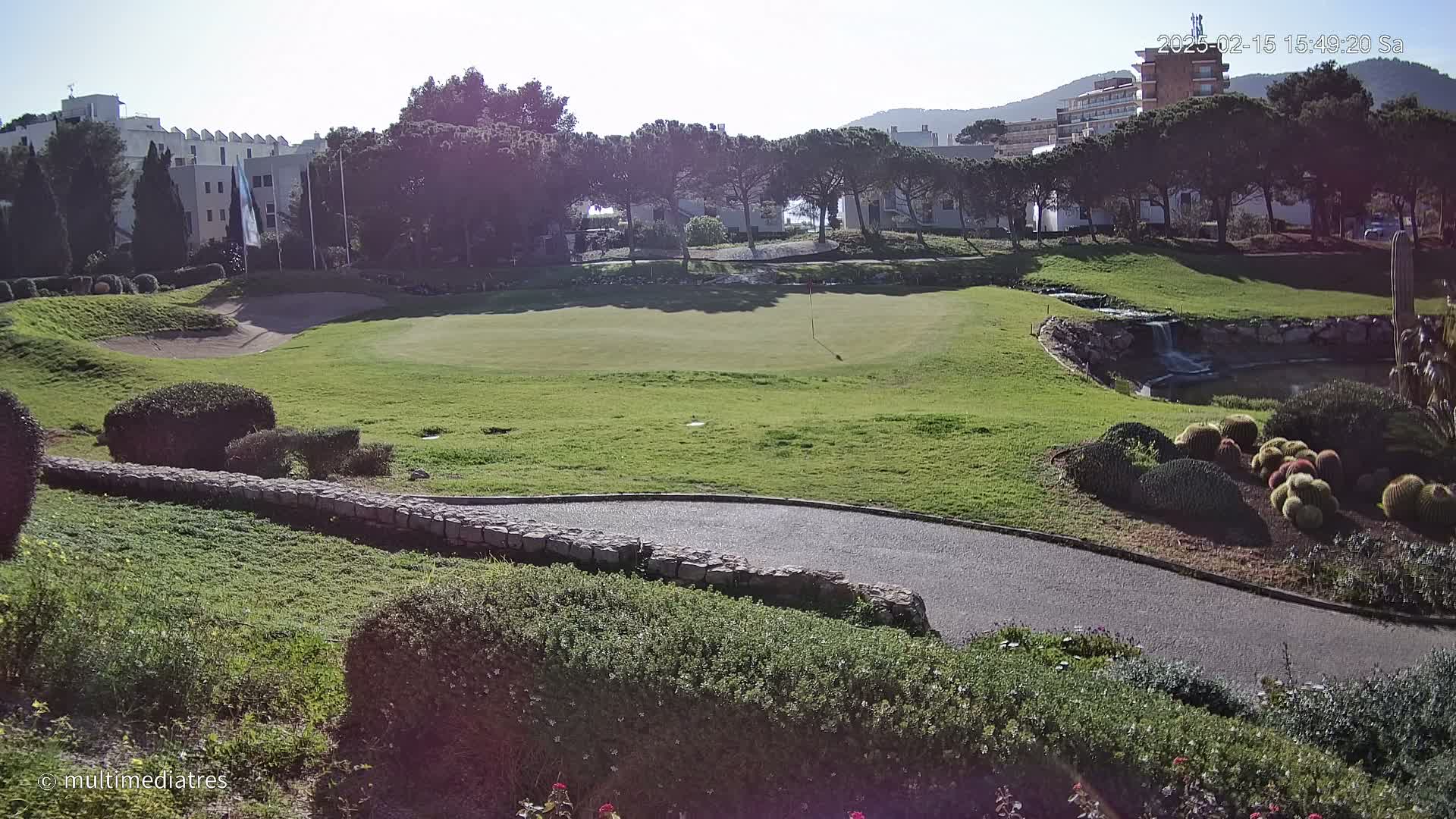 A sunny golf course with a green, sand trap, a small waterfall, and a variety of landscaping features including cacti, is visible, with buildings and trees in the background.
