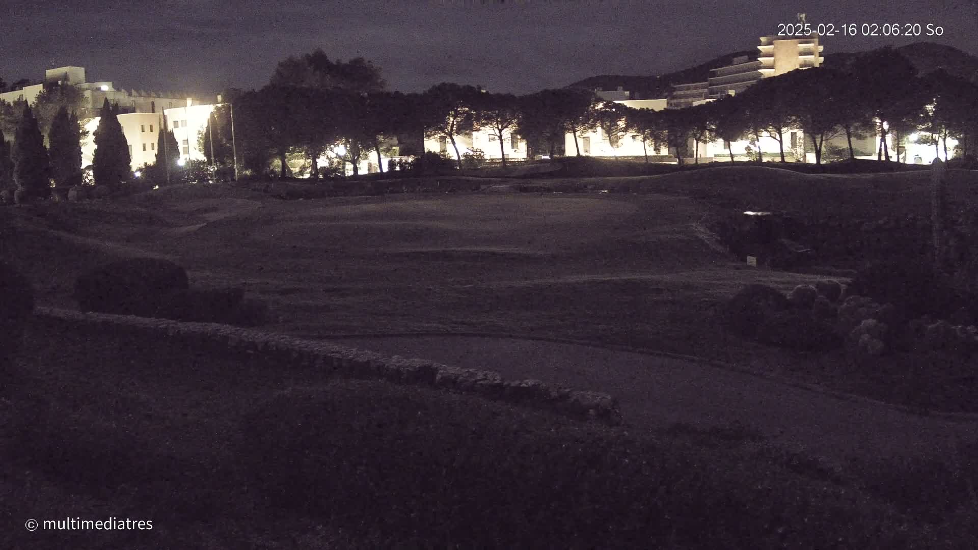 A nighttime view of a grassy field with buildings and trees illuminated in the background under a dark sky.