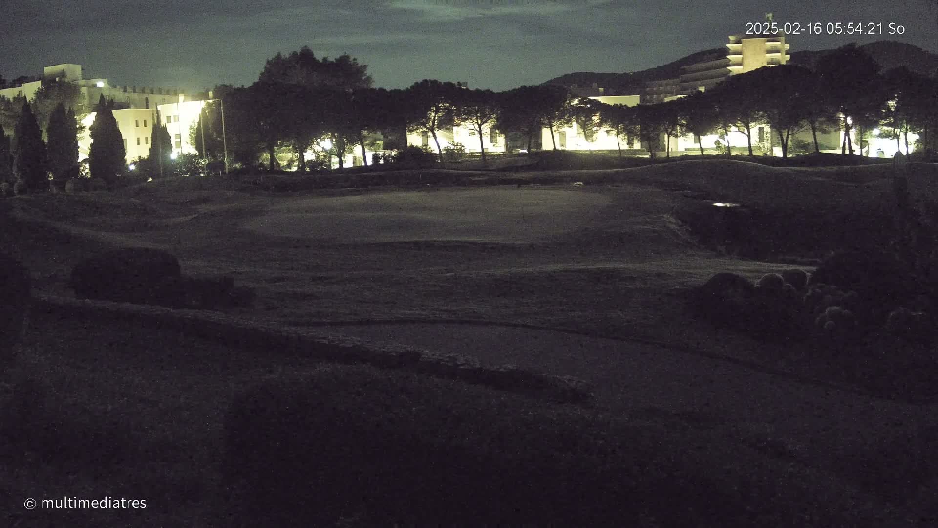 A nighttime view of a grassy area with buildings and trees illuminated in the background under a dark sky.