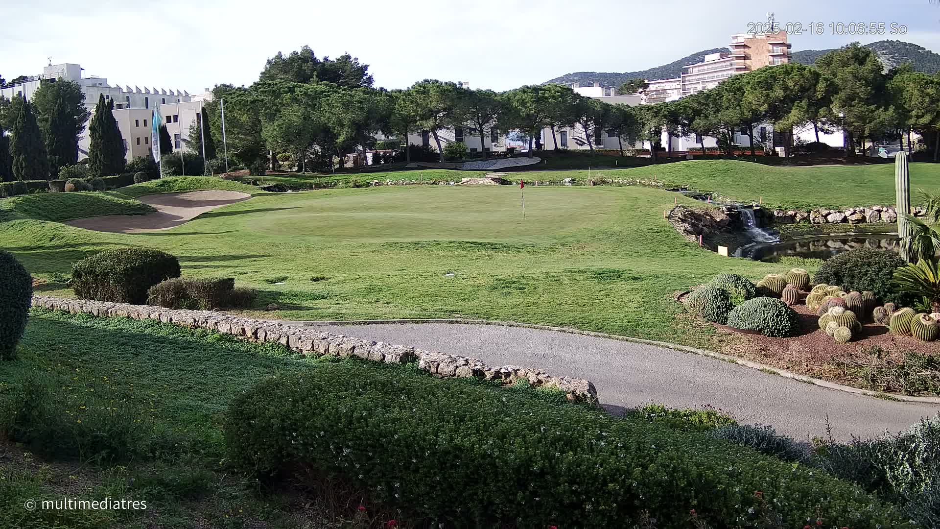 A golf course green with a sand trap, a small waterfall, and a pond, all situated near buildings and a line of trees under a clear sky.