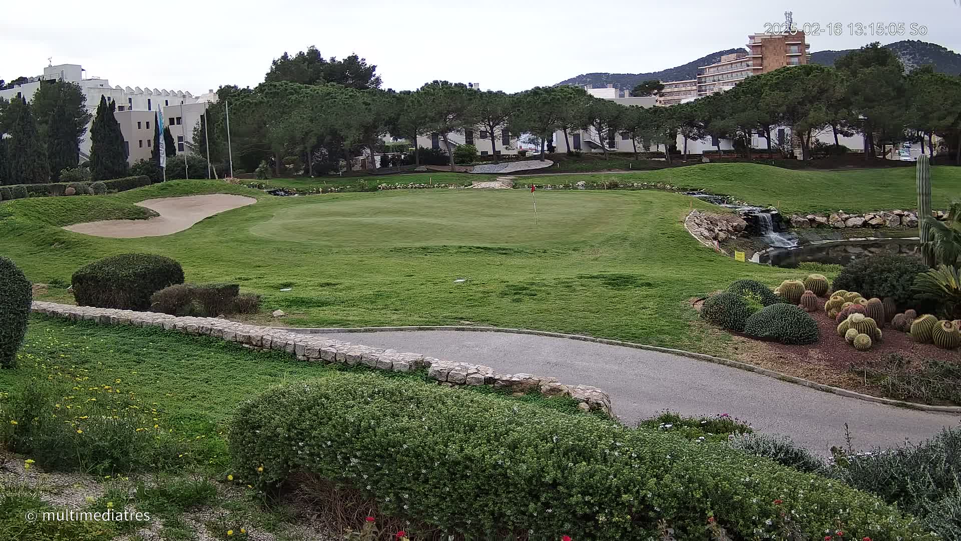 A golf course green with a sand trap, a small waterfall, and a variety of shrubs and cacti, under an overcast sky.