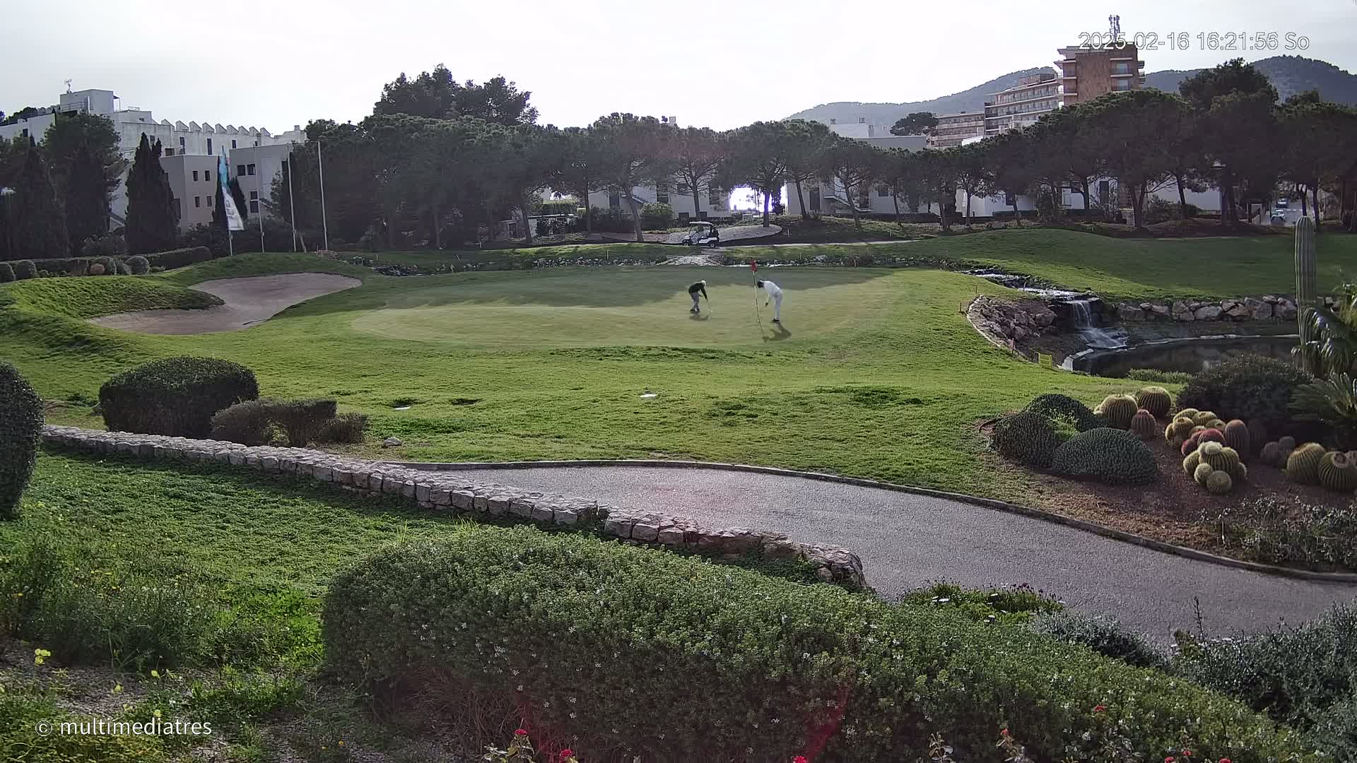 Two golfers putt on a green overlooking a small pond and a resort building under a partly sunny sky.