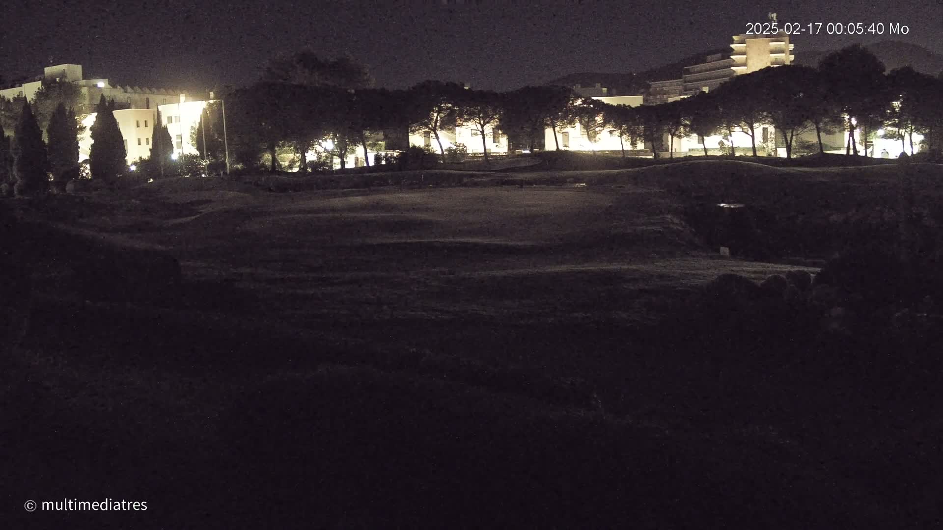A nighttime view of a dark, grassy area with a row of illuminated buildings and trees behind it under a clear sky.