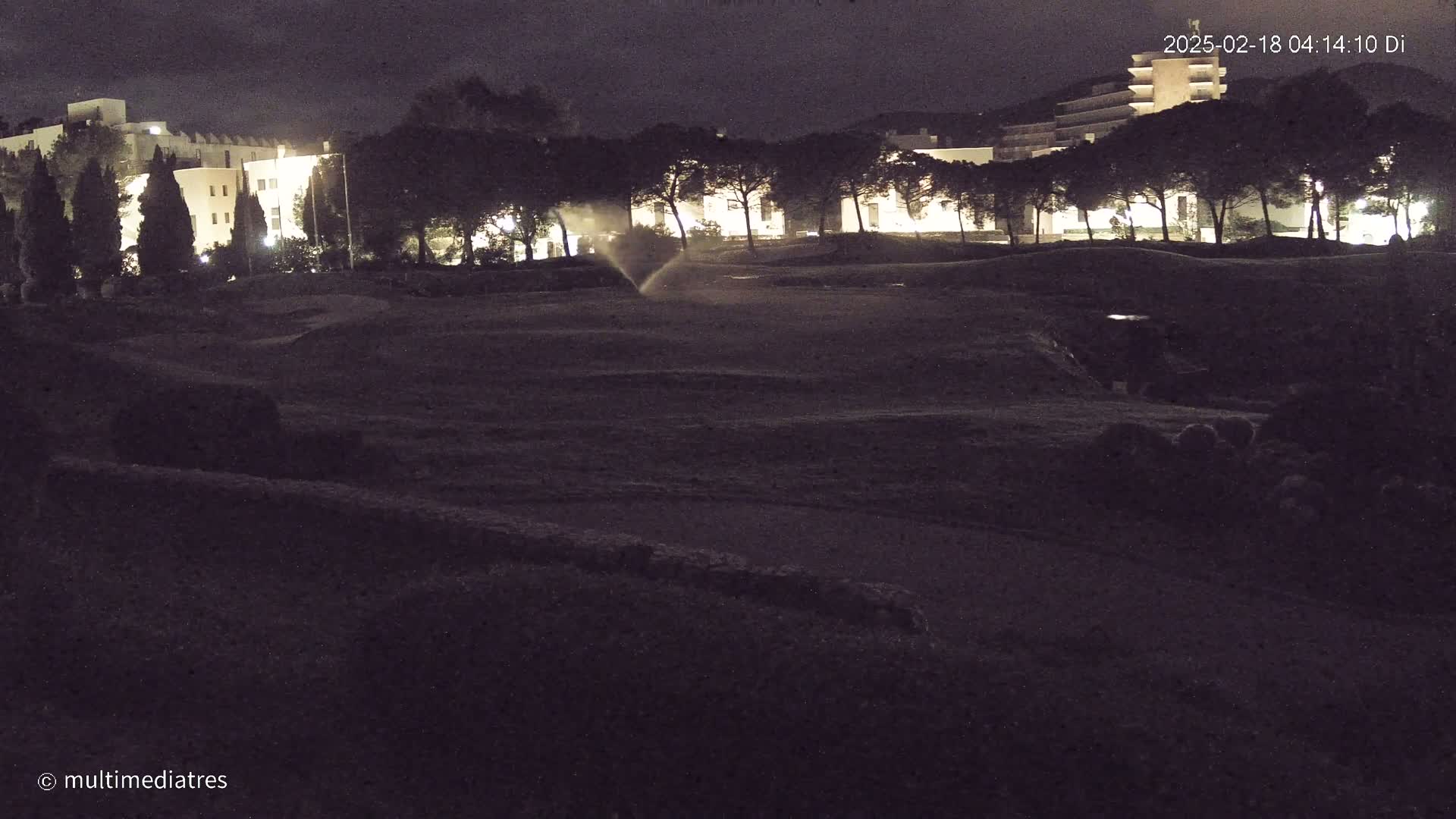A nighttime scene shows a sprinkler irrigating a dark, grassy area in front of a row of illuminated buildings and trees.