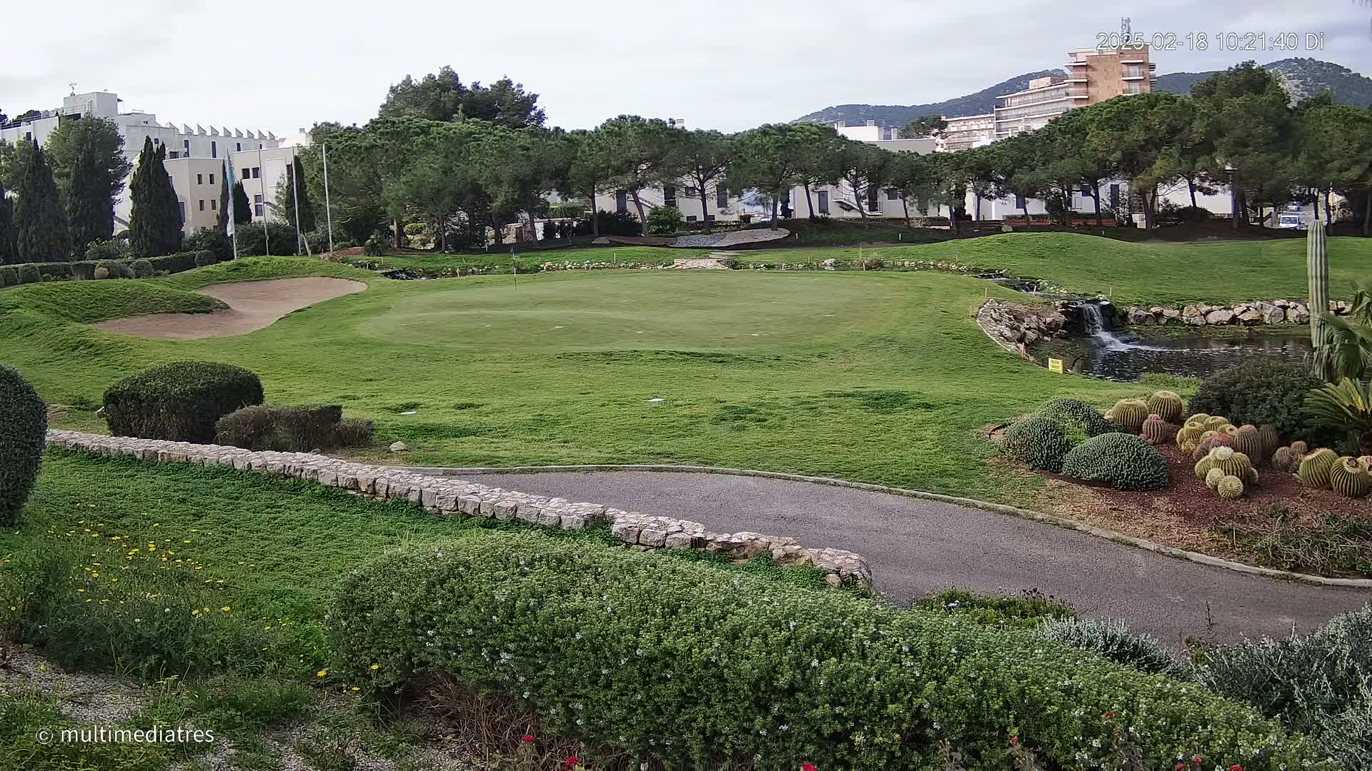 A golf course green with a sand trap, a small pond, and a low stone wall, under an overcast sky, is surrounded by lush green landscaping and buildings in the background.