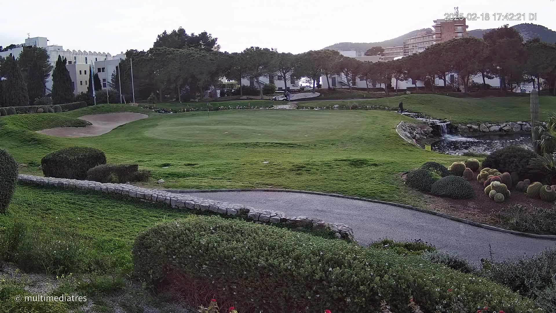 A golf course with a putting green, a small pond with a waterfall, and a collection of cacti, situated near buildings and trees under a cloudy sky.