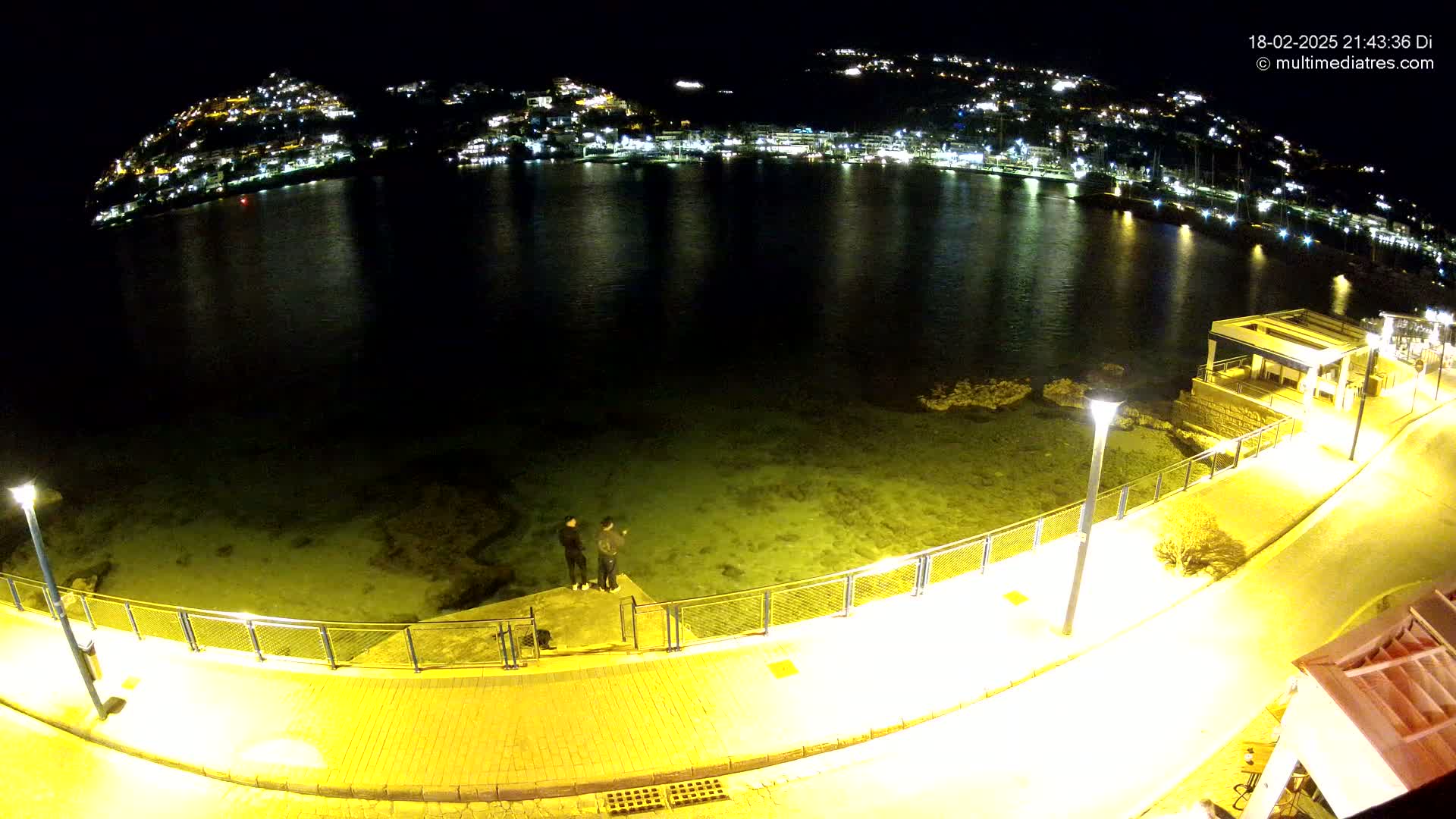 A nighttime view of a calm, clear body of water reflecting the lights of a town on a hillside, with a paved walkway and two people standing near the water's edge.