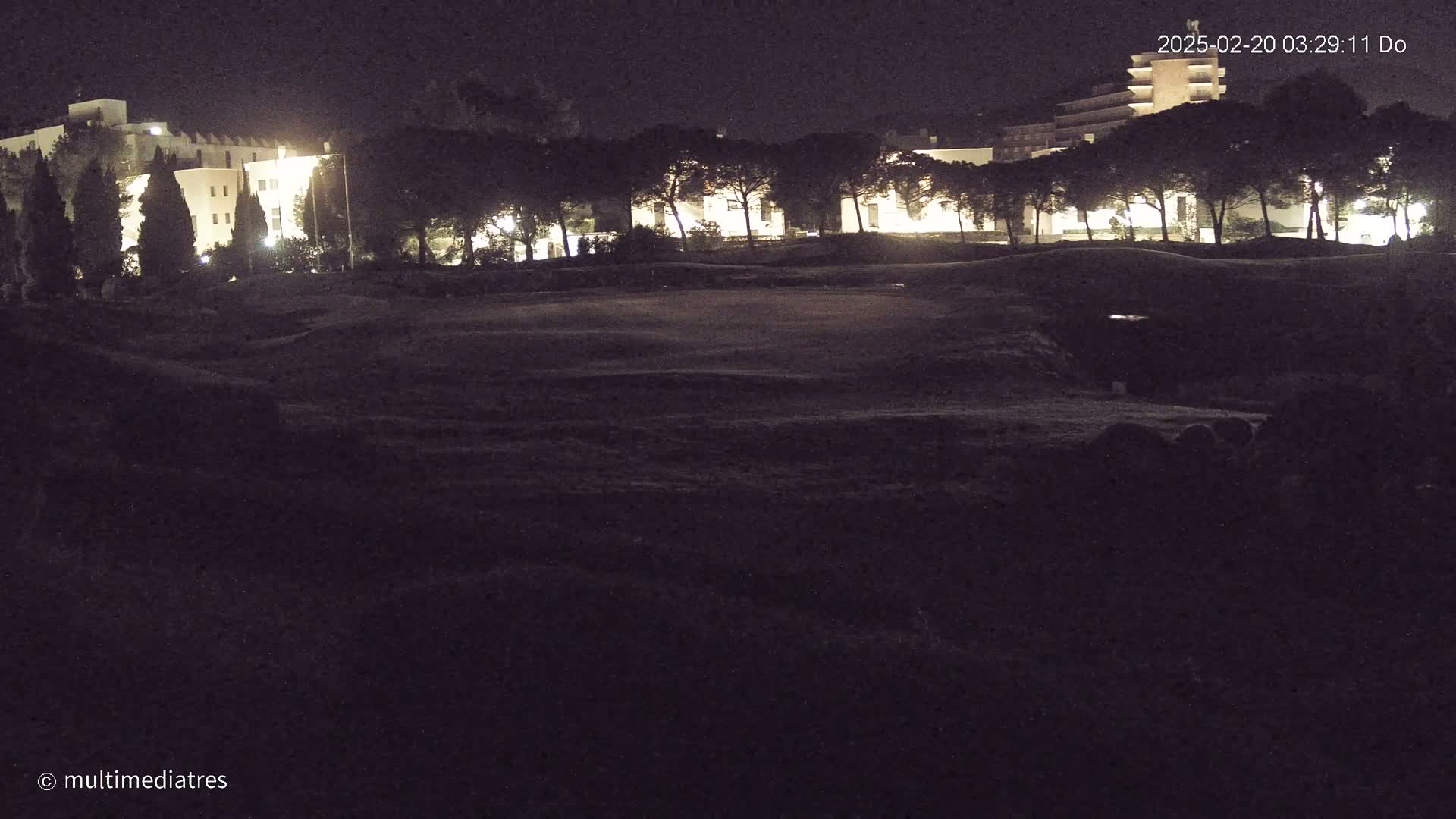 At night, a dark, grassy area stretches before illuminated buildings and a line of trees.