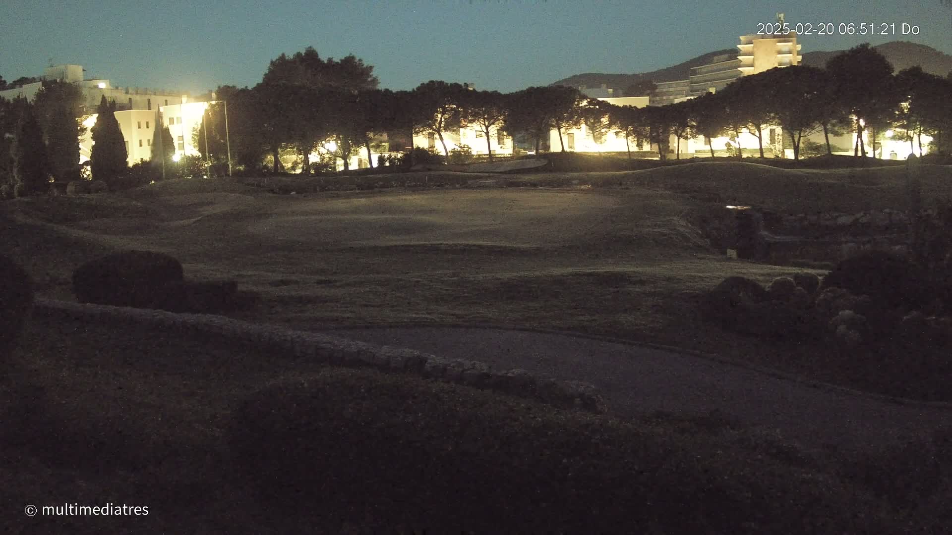 A nighttime view of a grassy area with illuminated buildings and trees in the background under a dark sky.