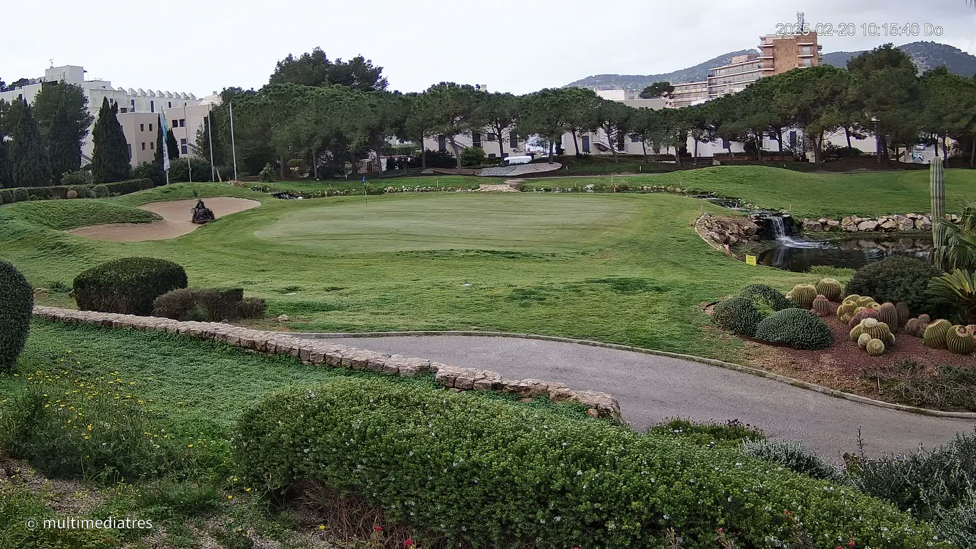 A golf course with a green, sand trap, small pond with a waterfall, and a variety of shrubs and cacti under an overcast sky.