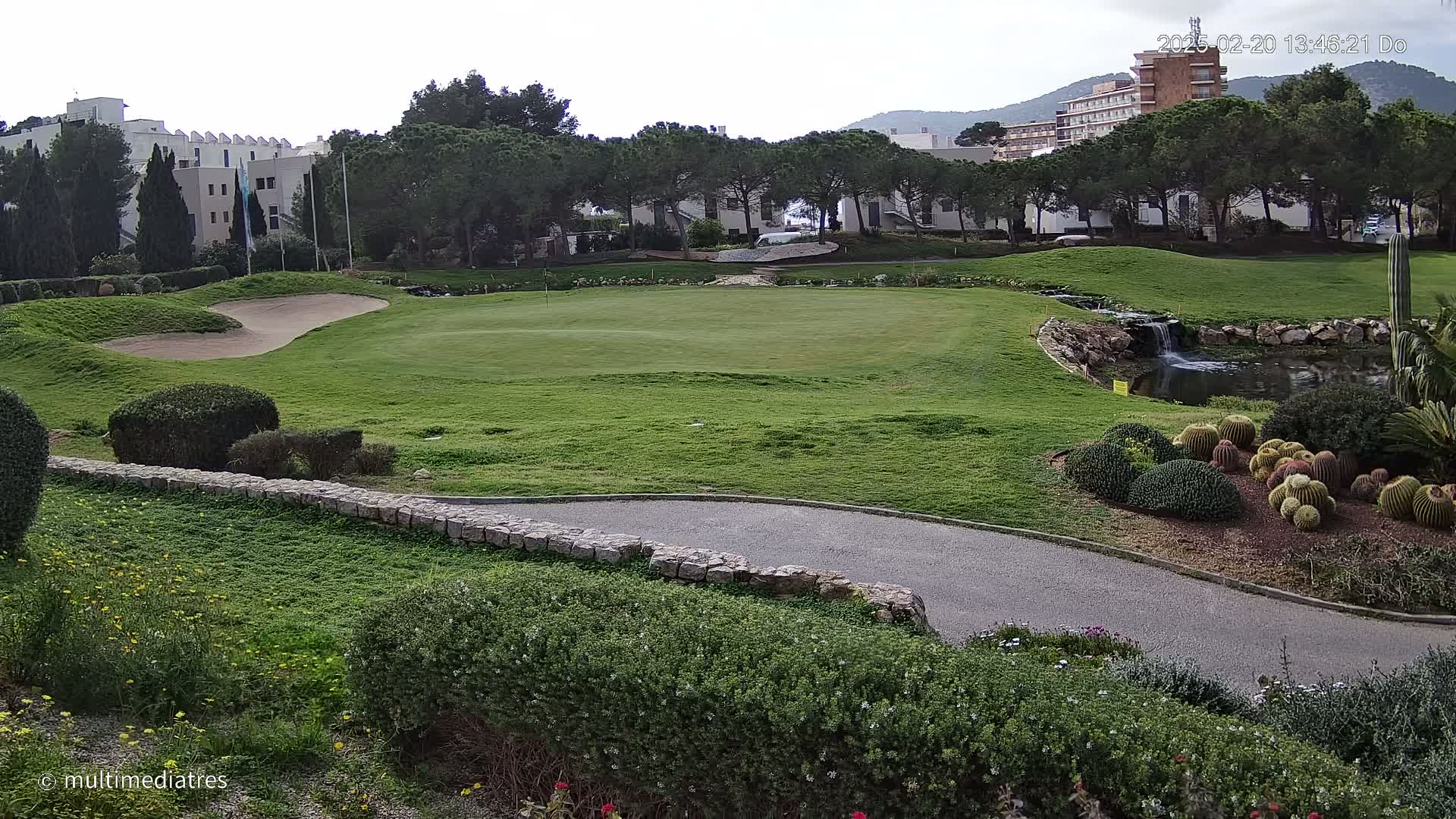 A golf course green with a sand trap, a small waterfall and pond, and a variety of cacti and shrubs, under an overcast sky.